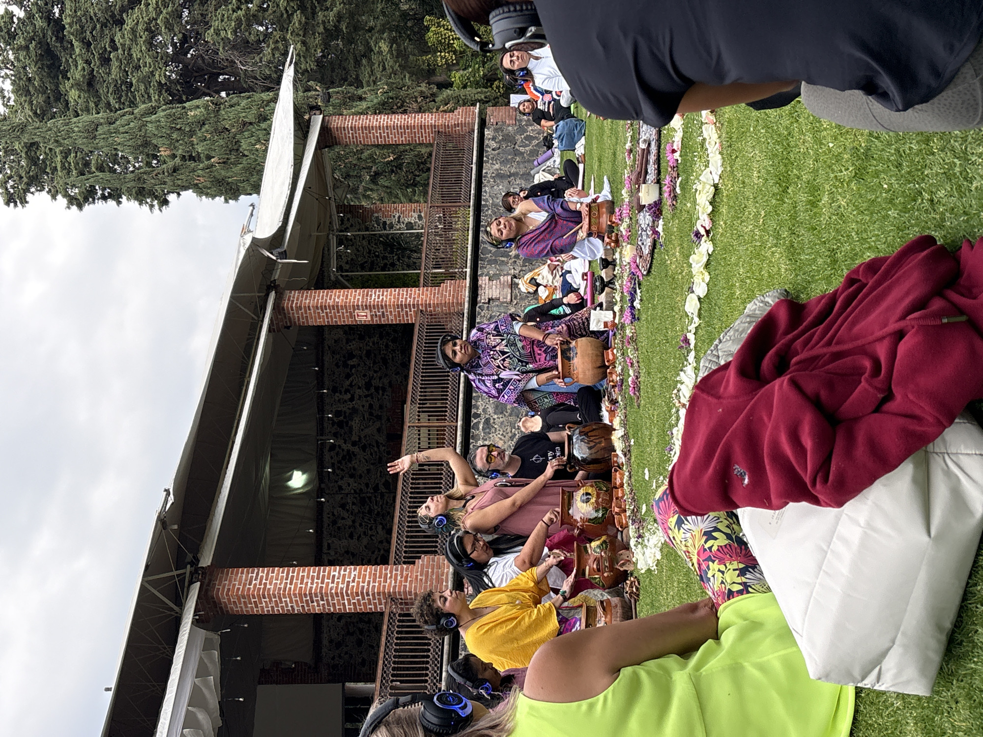 A wide shot captures a group of people seated on green grass in Santa Ana Acozautla, Mexico. They are arranged in a semi-circular formation facing a building with brick supports. The group of people are wearing headphones. Many of the people are seated on decorative stools, surrounding a circle of flowers. One woman is extending her arm. The grassy area is flanked by trees in the background, beneath a cloudy sky.