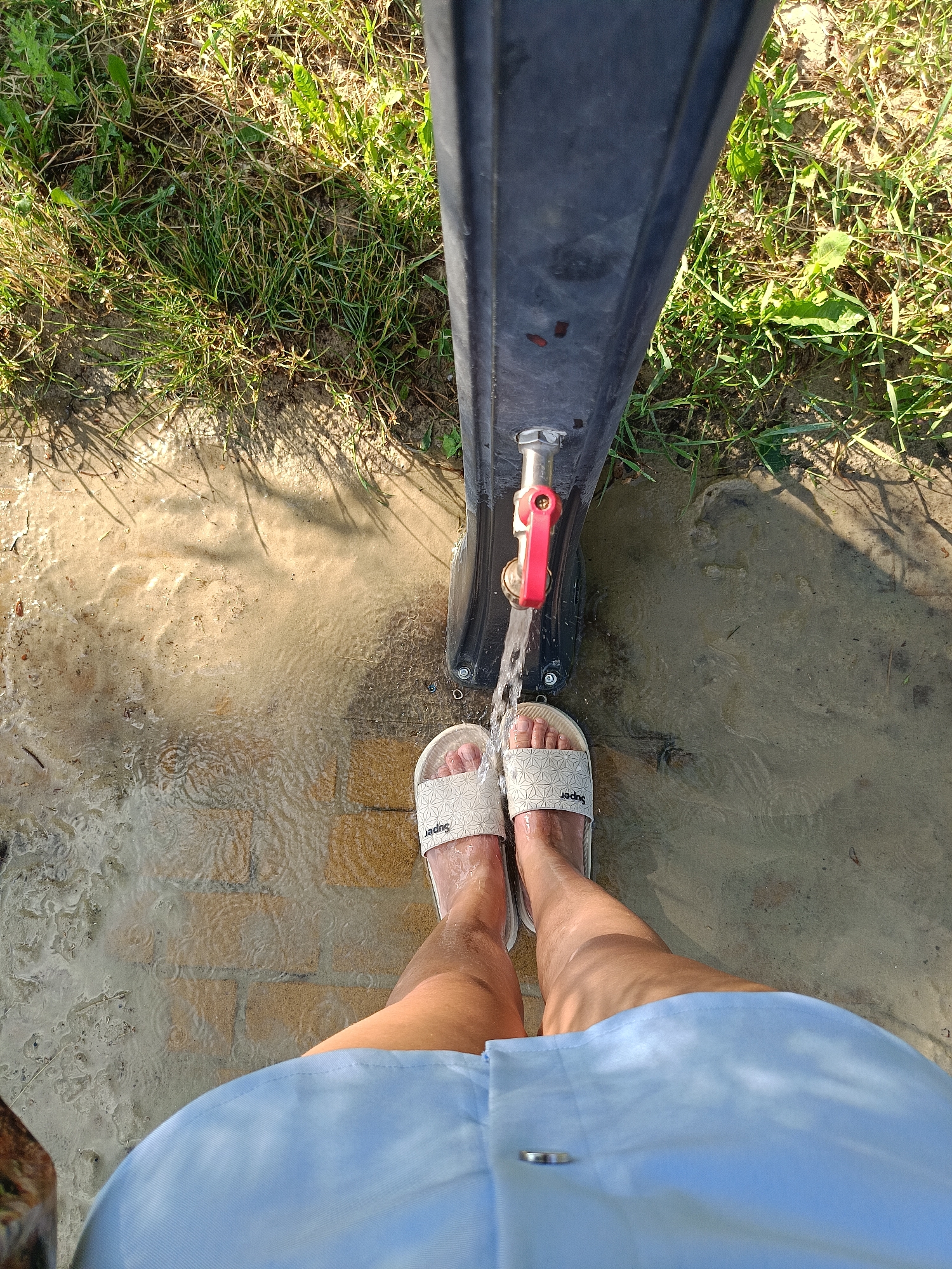 The image depicts a ground-level view of a person's legs and feet wearing white slides with the word "Super" on them. The person is standing on a wet, paved surface, likely a pathway or patio, with water flowing from a spigot attached to a dark, vertical structure. The water creates ripples and puddles on the ground. Green grass and some dry earth are visible in the background, with dappled sunlight casting shadows. The scene appears to be outdoors, possibly in a garden or yard area. The location of Gubkin, Russia is not discernible from the visual information provided in the image.