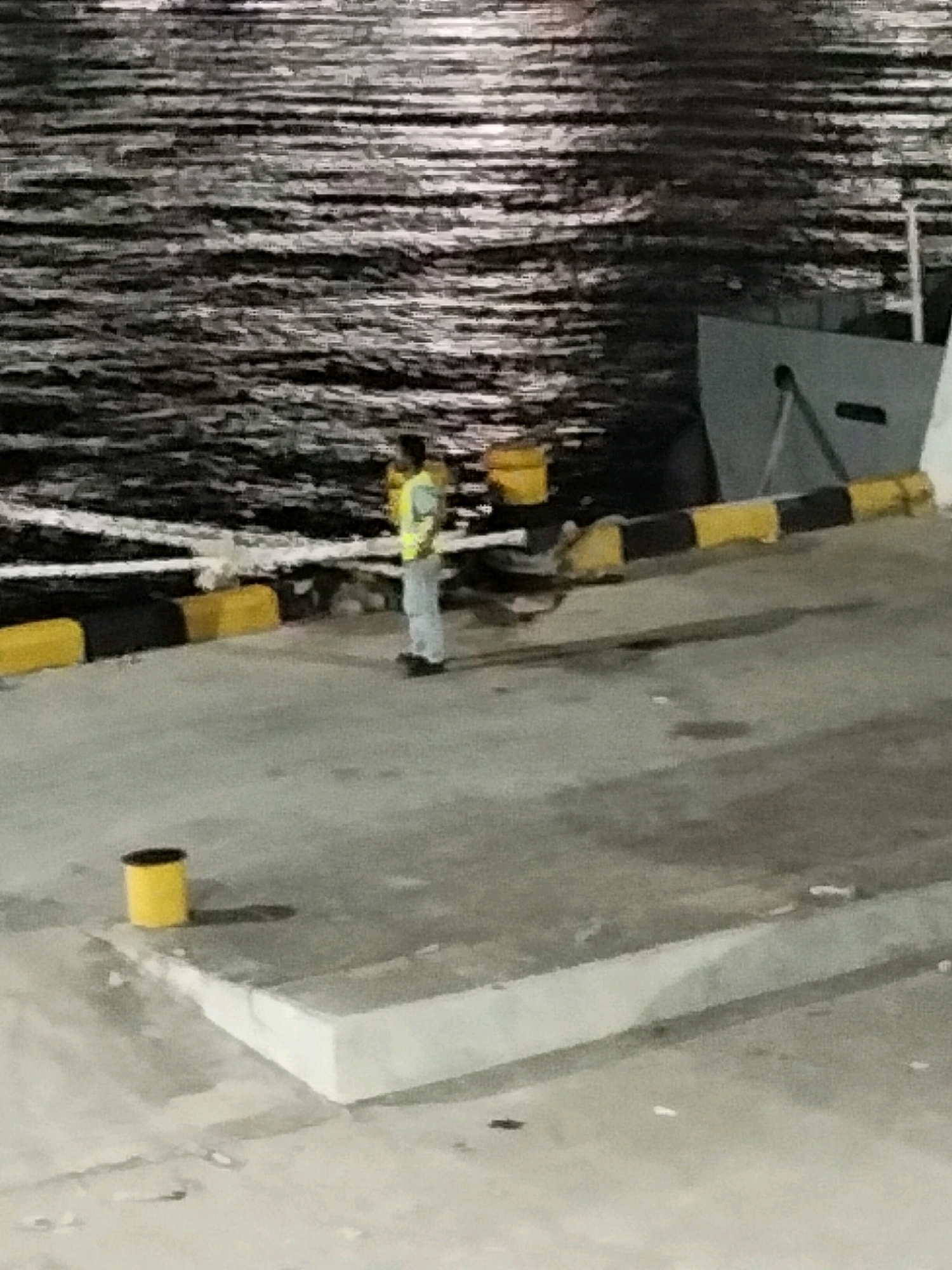 An individual wearing a high-visibility vest and light-colored pants stands on a concrete pier. The pier is bordered by a yellow and black striped barrier. To the left of the individual, a yellow cylindrical object, possibly a bin, is positioned on the concrete. In the background, a dark, textured body of water with reflections is visible, and a portion of a grey structure, likely a ship or building, is present. The scene appears to be a port or dock area.