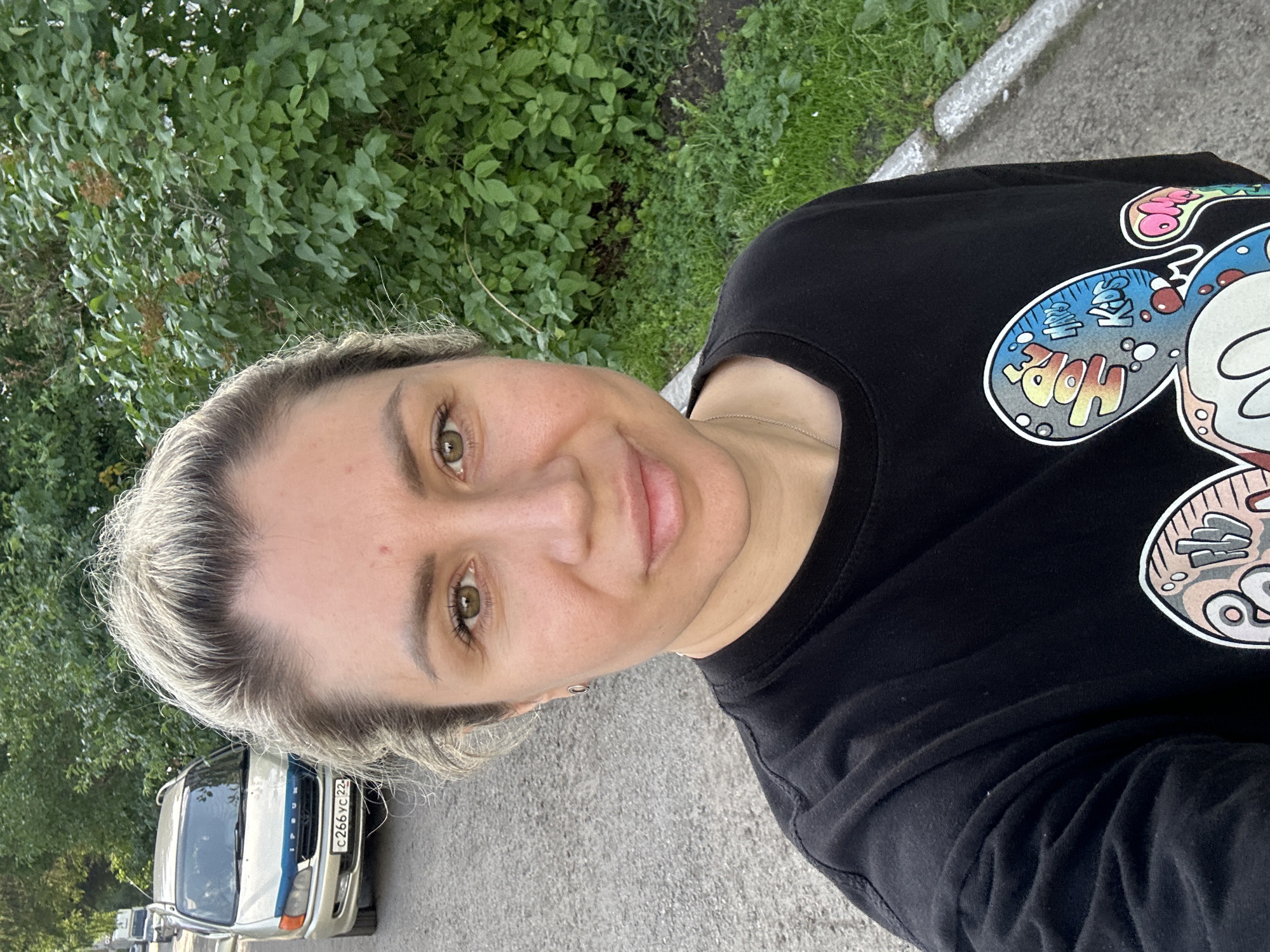 A woman with blonde hair pulled back is taking a selfie outdoors. She is wearing a black t-shirt with a graphic design of Mickey Mouse on it. Behind her, a silver minivan with a blue accent on the hood is parked on a paved road. The road is lined with grass and bushes. The license plate of the minivan is visible and reads "C 266 YC 22". The background suggests a residential area in Novoaltaysk, Russia, possibly near a park or green space. The lighting indicates it is daytime, with a slightly overcast sky.