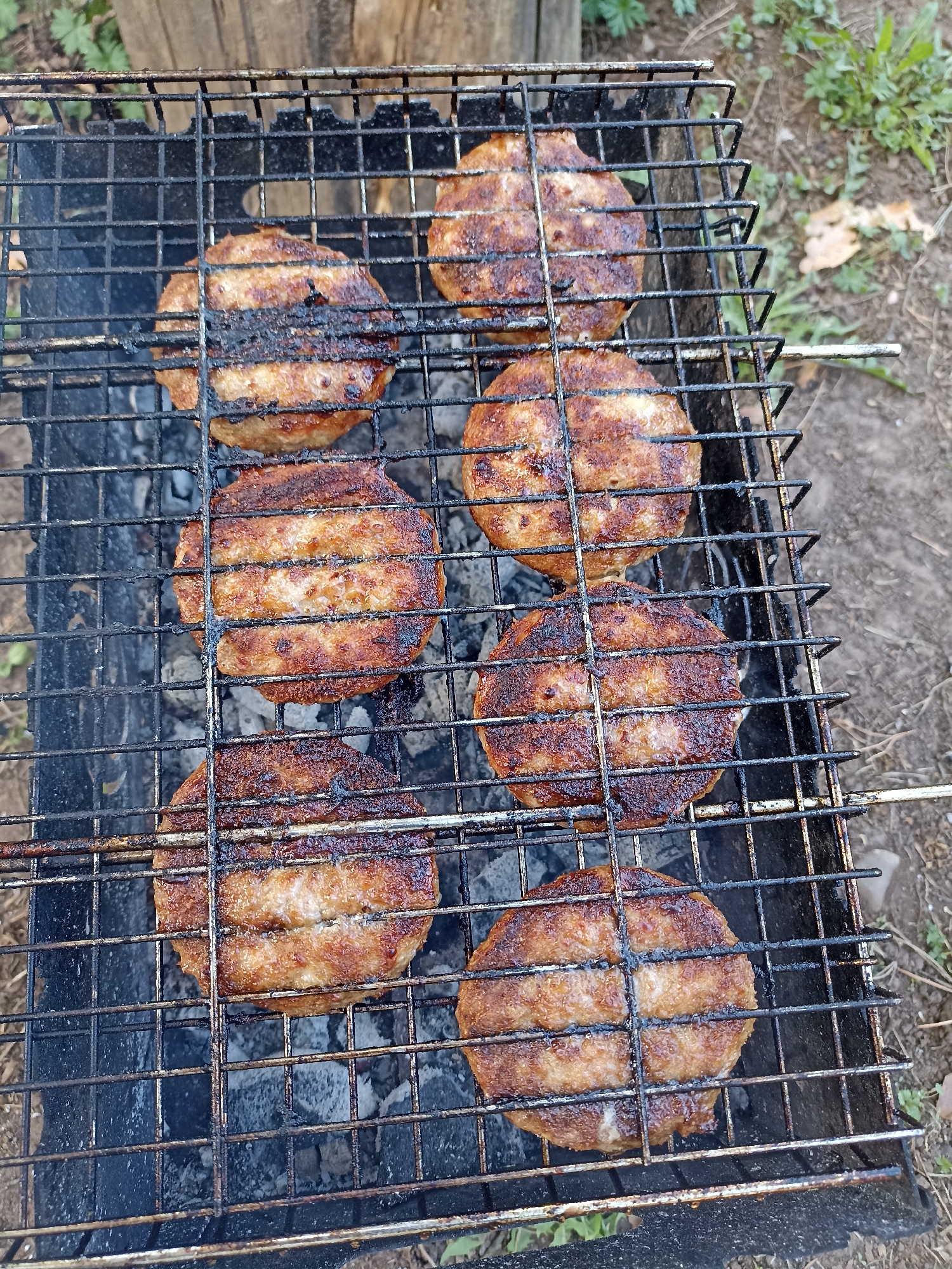 The image shows six round, cooked burger patties on a black metal grill over a charcoal-filled grill pit, likely outdoors. The patties have grill marks. Background details suggest an outdoor setting. No specific evidence indicates the location is Chaikovsky, Russia.