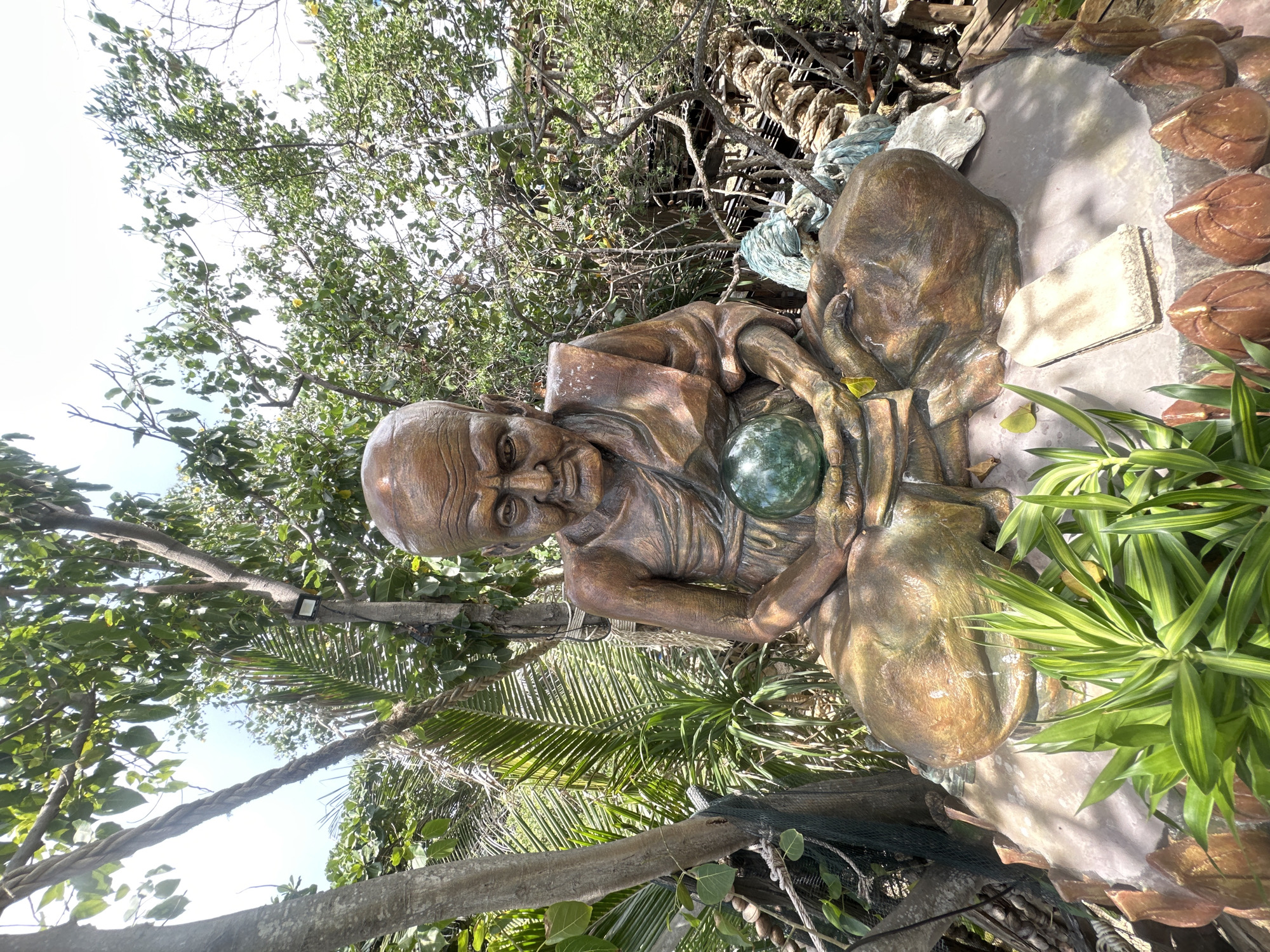 A statue of an elderly monk is seated cross-legged on a rock in a garden environment. The statue is made of weathered material, giving it a bronze appearance, and depicts the monk with a bald head, wrinkled face, and wearing a saffron robe. The monk's hands are cupped together, holding a smooth, green, spherical object, possibly a large marble or glass orb. The statue is situated on a raised platform adorned with decorative elements, including what appear to be carved wooden or ceramic teardrop shapes at the base. Lush green foliage, including palm fronds and other leafy plants, surrounds the statue. In the background, trees with dense green leaves create a natural canopy, and a clear sky is visible above. The setting suggests an outdoor location, potentially a temple or a garden display, in ตำบลเกาะพะงัน, Thailand.