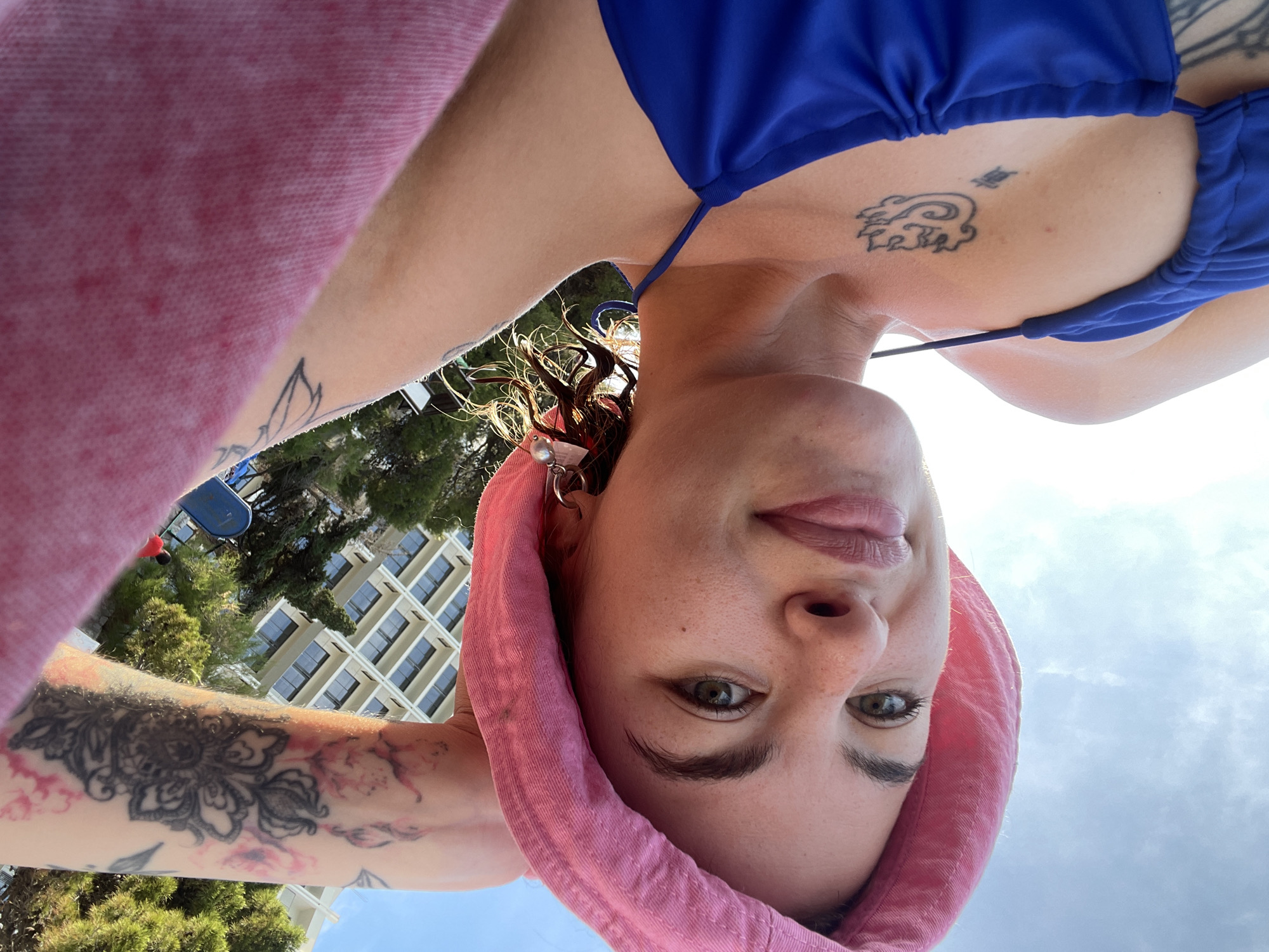 A woman is looking at the camera, head upside down, while outside in what appears to be a vacation setting. She is wearing a blue bikini top and has tattoos on her chest and arm. She has a pink towel wrapped around her head. In the background, there is a building with many windows and some trees, suggesting an urban or resort area. Based on the context of Rafailovići, Montenegro, it is plausible that this is a hotel or apartment building near the coast. There is blue sky visible, implying daytime.