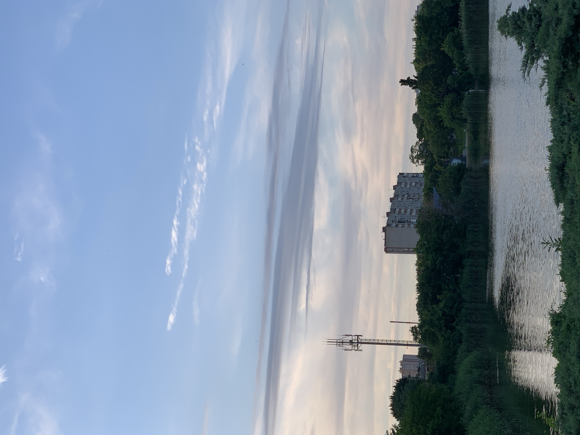 The image shows a body of water with a dense green shoreline. Beyond the shoreline, there are trees, buildings, and a telecommunications tower. The sky above is partly cloudy, with streaks of white and gray clouds against a blue background. The overall scene depicts an urban or suburban environment with elements of nature. The water is calm, reflecting the sky and surrounding greenery. The buildings are of varying heights and architectural styles, suggesting a mixed-use area. The telecommunications tower is tall and slender, with visible antennas. The lighting suggests it might be late afternoon or early evening.