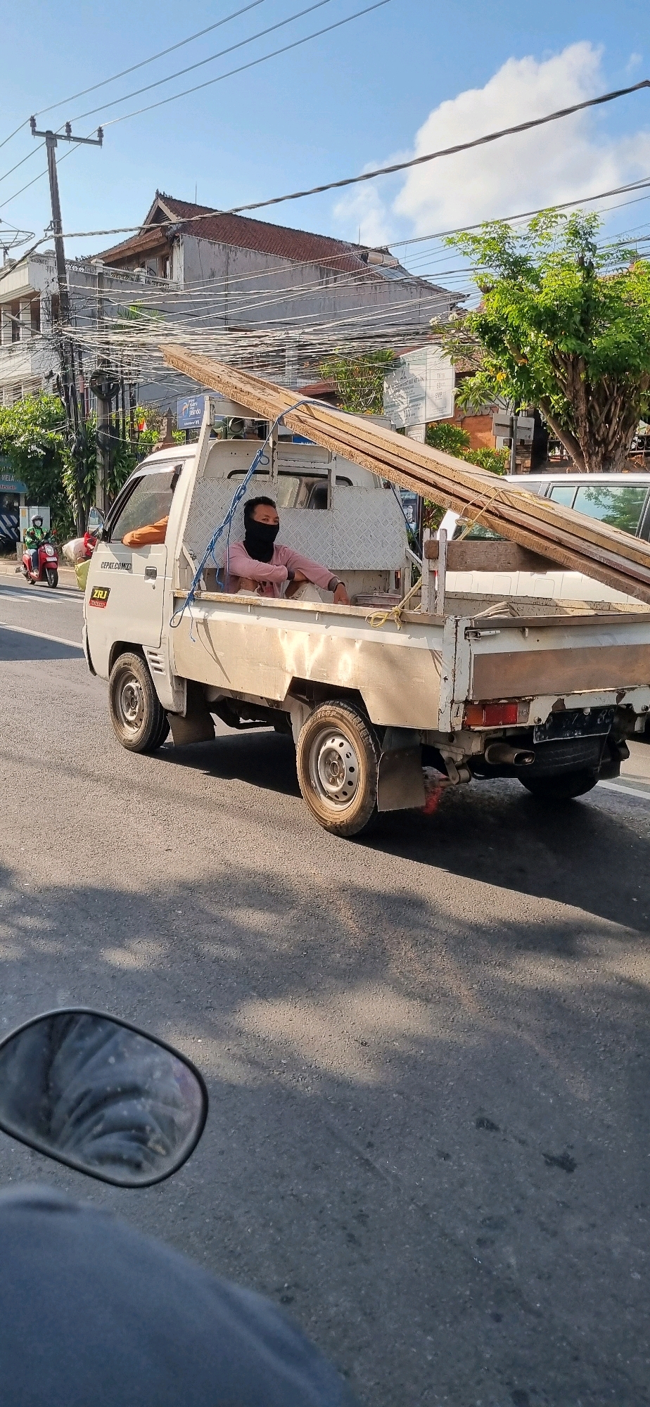 A white pickup truck is traveling on a street in Denpasar, Indonesia. The truck is carrying several long wooden planks stacked in its bed. A person wearing a pink shirt and a black face covering is sitting in the passenger seat of the truck, looking to the right. The truck is on a paved road with some shadows from trees on the right side of the image. In the background, there are buildings, trees, and a complex network of overhead electrical wires. A red scooter with a rider wearing a green helmet is visible in the distance on the left side of the road.