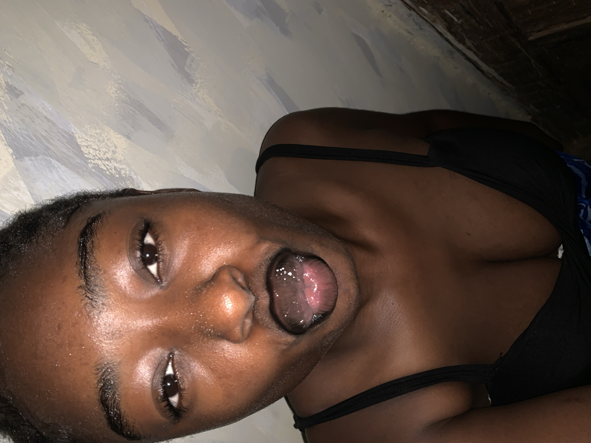 The image shows a close-up, head-and-shoulders shot of a young Black woman. She is looking directly at the camera with her mouth open, and her tongue is visible. Her skin is dark and appears moist, possibly from sweat or water. She is wearing a black bra or camisole with thin straps. The background is a textured wall with a pattern of light and dark strokes, likely painted. The lighting is dim, casting shadows and highlighting the woman's features. There is no indication of the specific location within Rafin Par, Nigeria.