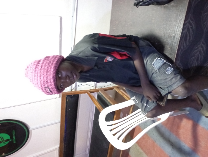 The image depicts an indoor scene featuring a person seated on a white plastic chair. The person, appearing to be male with a dark skin tone, is wearing a pink knitted beanie hat, a dark-colored short-sleeved collared shirt (possibly black or dark blue) with red detailing on the right shoulder/sleeve and a logo or emblem on the left chest, and ripped light-wash denim shorts. They are looking directly towards the camera with a neutral expression.
The person's left hand is resting on their lap, seemingly holding a small, rectangular object like a phone or wallet. Their bare feet are visible, resting on a patterned rug or mat with reddish-brown and lighter striped sections. To the right of the person, there is a dark, possibly wooden, surface or table, on which a crumpled black plastic bag is placed.
In the background, a white wall is visible. To the left of the person, an oval-shaped, dark green sign or plaque with a white emblem and the text "PIXLAM MEDIA" is mounted on the wall. A wooden-framed structure, possibly part of a piece of furniture or a divider, is also visible behind the person. The lighting in the room appears bright and even, suggesting an interior space that is well-illuminated. The image is presented rotated 90 degrees clockwise from its likely original orientation.
