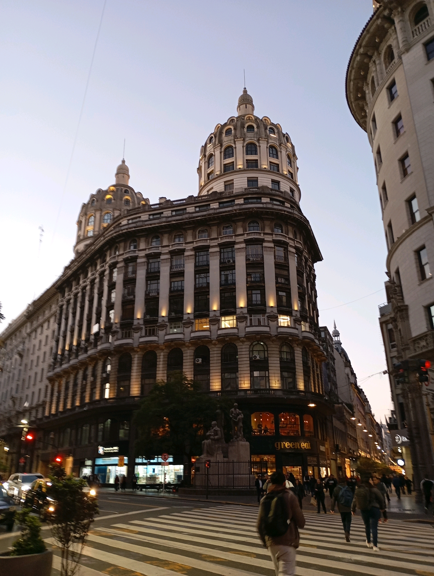 The image depicts a street scene in Buenos Aires, Argentina, likely at dusk given the lighting and sky. Dominating the frame is a large, ornate building with multiple stories, classical architectural elements, and a prominent dome structure topped with a finial. Another curved building is partially visible on the right, also featuring classical design. The street below is lined with more buildings, some illuminated from within. A pedestrian crossing is clearly marked with white stripes, and several individuals are walking across it. There are vehicles on the street, including cars and potentially a bus, with their headlights on. Traffic lights are visible, one of which is red. Trees are present along the sidewalk, and streetlights are on, casting warm light. A sign for "green eat" is visible on one of the ground-floor businesses. The overall impression is of a busy urban environment with significant architectural heritage.