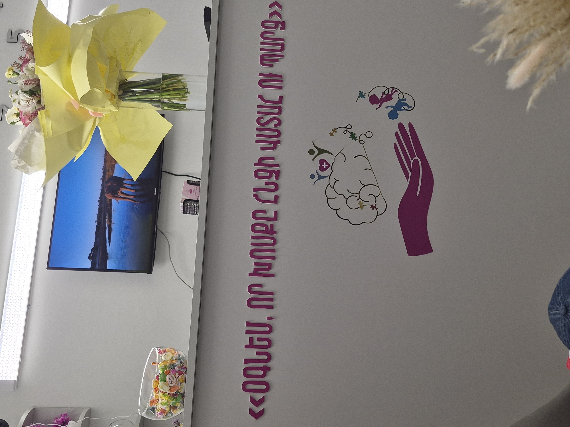 The image captures an indoor scene, likely a modern office or clinic in Yerevan, Armenia, characterized by light-colored walls and contemporary decor. On the left side, a bouquet of flowers wrapped in yellow paper is placed in a tall glass vase. Below the bouquet, a flat-screen television is mounted on the wall, displaying an outdoor scene with a clear blue sky. A large mirror is positioned along the left edge of the frame, reflecting parts of the room, including a clear container filled with colorful decorative elements near the bottom.
The central and right portions of the wall feature distinctive graphic art and text. A vertical phrase written in Armenian with pink, three-dimensional letters reads from bottom to top: «ՕԳՆԵՄ, ՈՐ ԴՈՒՐՍԴ ԳԱՆ». This translates to "I WILL HELP, SO THEY COME OUT" or "I will help, so your inner self emerges."
Adjacent to the text, further to the right, is a stylized illustration depicting a purple-colored hand, palms upwards, beneath two outlined brains. Various small, colorful figures resembling babies or children, along with abstract shapes, appear to be emerging from or being gently held/guided by the hand and associated with the brains. This imagery, combined with the text, suggests a focus on development, mental well-being, or nurturing growth. A fluffy, light-colored decorative plant or feather arrangement is visible in the upper right corner, adding to the soft aesthetic of the space. The overall impression is one of a calm, supportive, and possibly therapeutic environment.