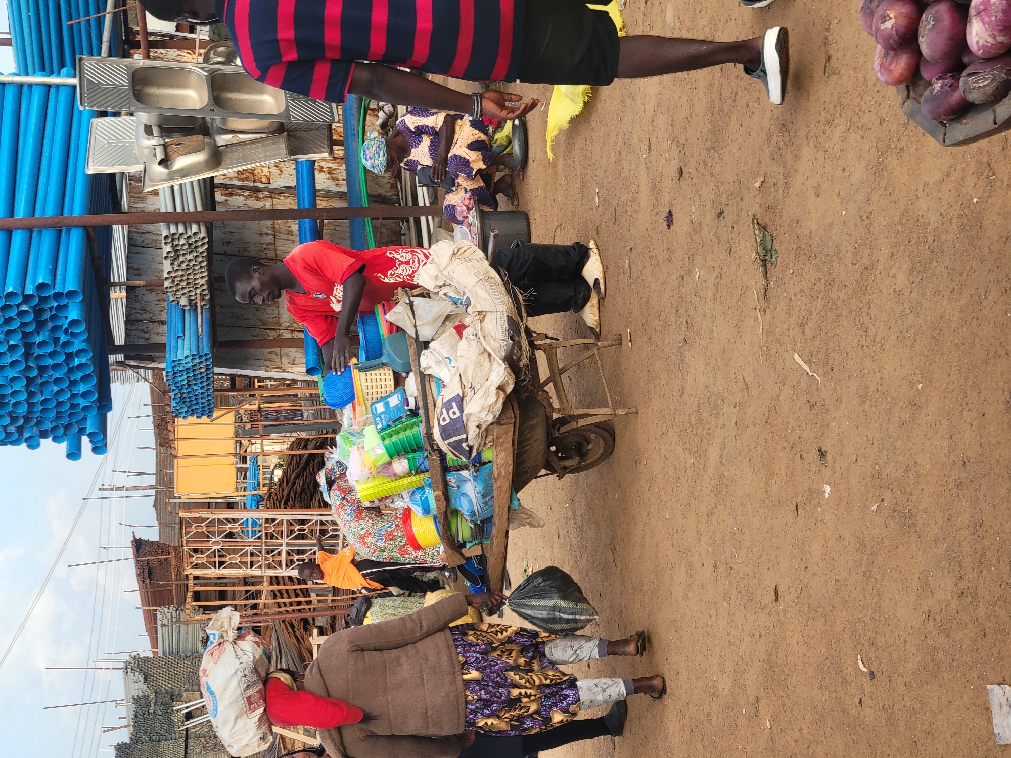 This image captures a bustling market scene in Gura Top, Nigeria. In the foreground, a person in a red shirt is sorting through a cart filled with colorful plastic goods, including basins, containers, and buckets. The cart is overflowing with various items, suggesting a lively trade. To the left of the cart, a stack of blue plastic pipes is prominently displayed. Behind the cart, several individuals are present. One person, dressed in a patterned fabric and a headscarf, appears to be resting or engaged in conversation. Another person, wearing an orange t-shirt, is also visible in the background.
In the upper portion of the image, several stainless steel sinks are stacked, indicating a hardware or plumbing supply section of the market. The ground is a dusty, sandy earth, typical of many outdoor markets. The overall atmosphere suggests a busy commercial environment, likely during daylight hours, though no specific time can be determined. The presence of various goods, from plumbing supplies to household items, points to a diverse market catering to different needs. There is no visible text in the image.