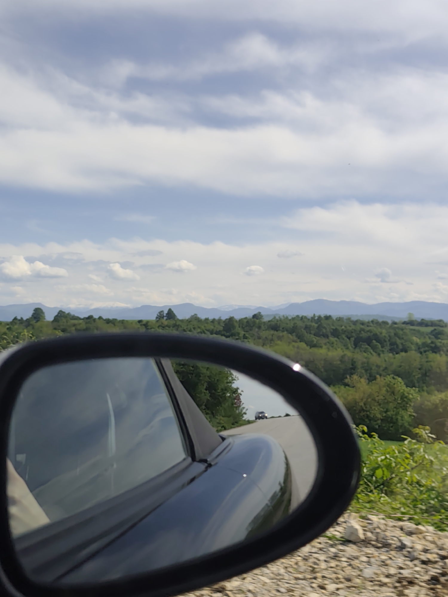 The image shows a view from the side mirror of a vehicle, capturing the scene behind it. The mirror reflects a stretch of road with another vehicle visible in the distance. The environment beyond the road is characterized by lush greenery, including trees and vegetation. In the background, a mountain range is visible. The sky is partially covered with clouds. The vehicle is on a gravel or dirt road. The city and specific location are unknown.