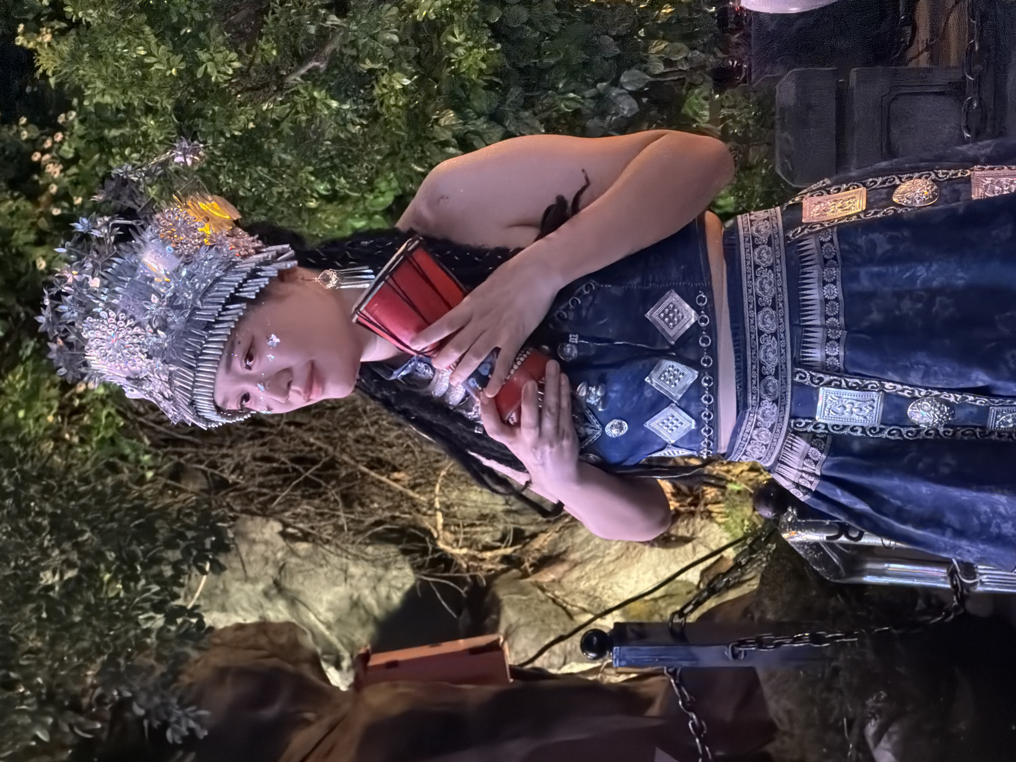 A person dressed in traditional attire stands in front of a lush green backdrop with rocky elements. The individual wears a large, ornate silver headdress adorned with intricate patterns and decorative elements. Their outfit consists of a dark blue, embellished crop top and a matching skirt. The clothing features silver diamond-shaped appliqués and decorative borders. The person holds a small red drum in their hands, with their fingers wrapped around it. They have long, dark braided hair and wear dangling earrings. Their face is adorned with subtle glittery makeup. The background suggests an outdoor or stage setting, possibly at an event or performance. The lighting is dim, with a focus on the individual. The location is the city of Yuzhong District, China.