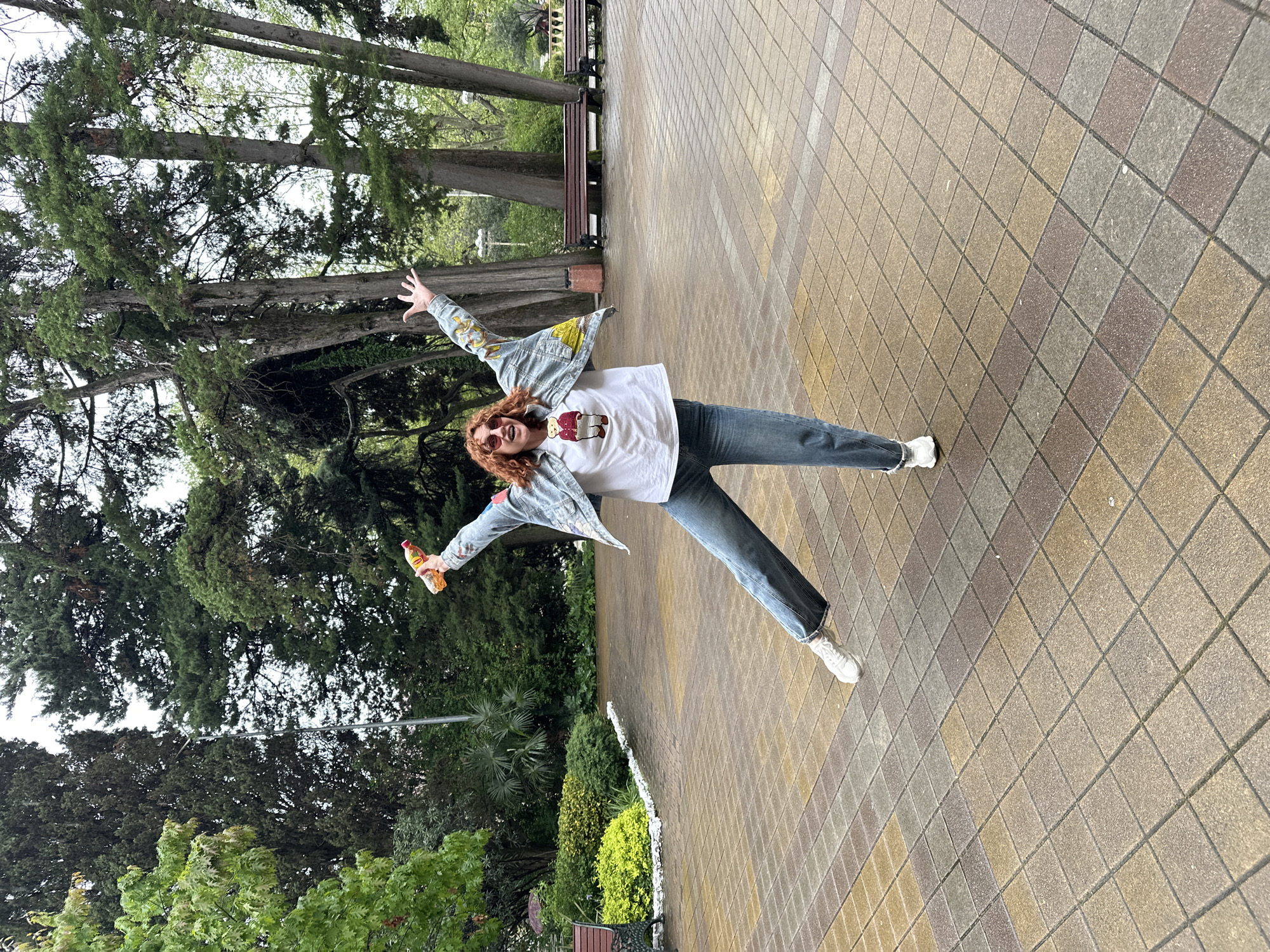 A person lies horizontally on a pedestrian walkway in a park setting. The person is wearing a white T-shirt with a graphic, jeans, a denim jacket with colorful designs, sunglasses, and white sneakers. They are holding a plastic bottle in their right hand, with arms outstretched in a "star" pose. The location is likely a park or public area in Sochi, Russia, as suggested by the vegetation, benches, and architecture style. The walkway is made of multi-colored square pavers. The environment includes trees, bushes, and park benches. The action is posing for a photograph.