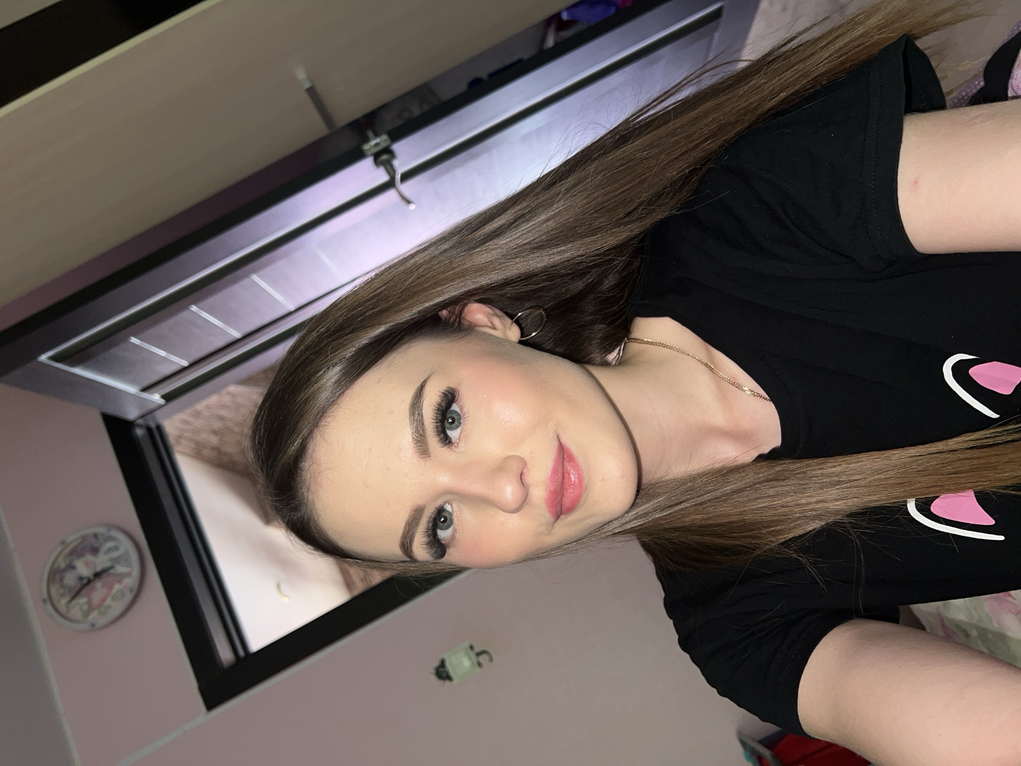 A young woman with light skin and long, light brown hair is posing for a selfie. She is wearing a black t-shirt with a pink and white design. She has blue eyes and pink lips, and is wearing hoop earrings and a gold necklace. Behind her is a partially visible closet or door, and a clock on a pink wall. Based on the presence of Russian letters in the image and assuming Noviy refers to a location in Russia, this image appears to be taken indoors in a residence.