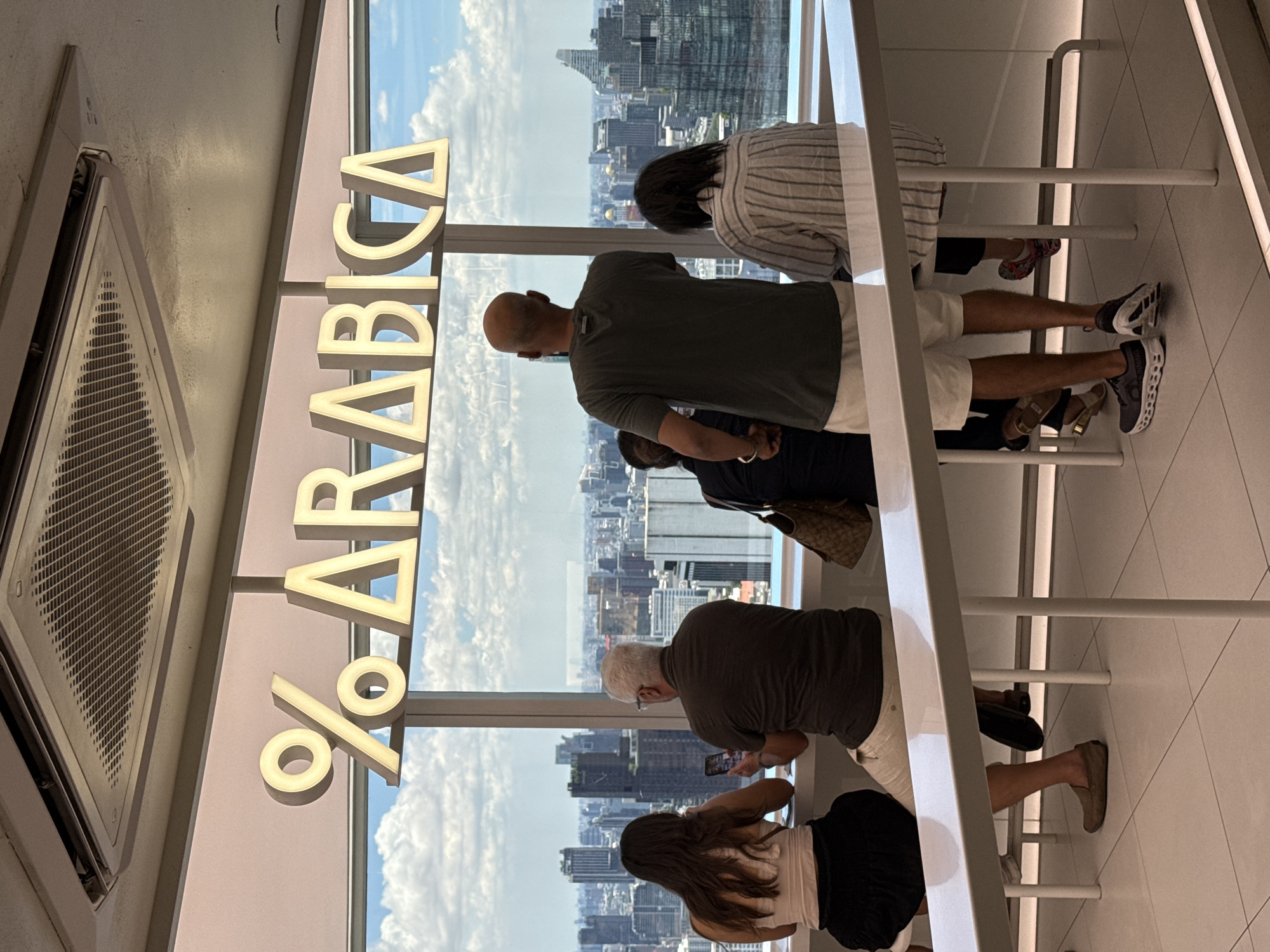 The image shows an elevated view from inside a structure overlooking Bangkok, Thailand. Four people are at a window, likely tourists viewing the cityscape. One person is taking a photo with their phone. The environment is bright and modern with clean lines. The logo "% ARABICA" is prominently displayed.