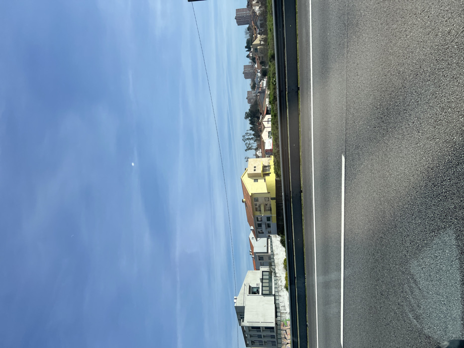 The image depicts a static daytime scene in Águas Santas, Portugal, captured from the perspective of a vehicle on a multi-lane roadway looking towards a residential area. In the immediate foreground, dark asphalt road pavement is visible, marked by a solid white lane line. A silver-colored metal guardrail spans horizontally across the frame, separating the road from the adjacent terrain. Beyond the guardrail, an upward-sloping embankment with sparse green vegetation leads to a cluster of residential and urban structures.

Prominently featured in the midground are two main buildings: to the left, a modern, light grey multi-story building with flat roofs and large, dark-paned windows; to its right, a multi-story house with a distinct yellow facade and a terracotta-tiled hip roof. Further in the background, numerous other residential buildings of varying sizes and rooflines are visible, interspersed with trees and culminating in a cluster of taller apartment buildings on the horizon. The sky is clear and blue with faint wisps of white clouds, and a bright, circular point of light, consistent with the sun or a strong reflection, is situated in the upper-center portion of the sky. A single dark utility wire stretches horizontally across the upper-midground, above the buildings. No individuals, vehicles other than the presumed source of the image, or explicit actions are discernible within the frame.