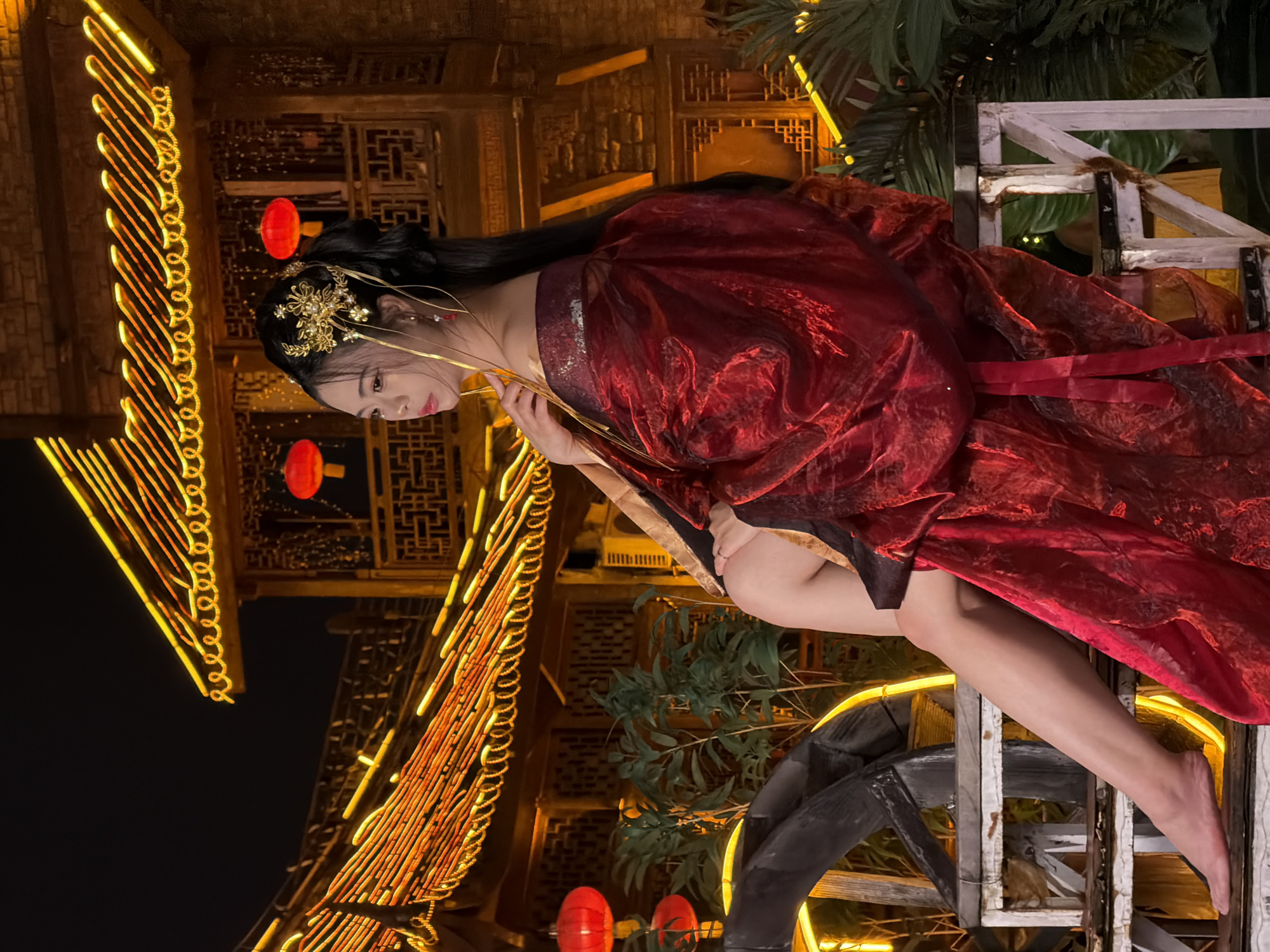 A young woman wearing a red, flowing, traditional Chinese dress and ornate gold hair accessories is seated on a white wooden structure at night. She is positioned on the right side of the frame, with her body turned to the left, her left leg extended and bare foot resting on a step. Her right arm is bent at the elbow, with her hand gently touching her chest.
Behind her, a traditional Chinese building with intricate wooden latticework and a curved roof is illuminated by warm, yellow string lights and several red lanterns. Lush green foliage and a dark, partially visible water wheel are also present in the foreground and midground, adding to the atmospheric setting. The scene appears to be in a well-lit, decorative outdoor area at night, likely a tourist or cultural site. The architectural style and decorations suggest a location in Yuzhong District, China, which is known for its historical buildings and traditional ambiance.
