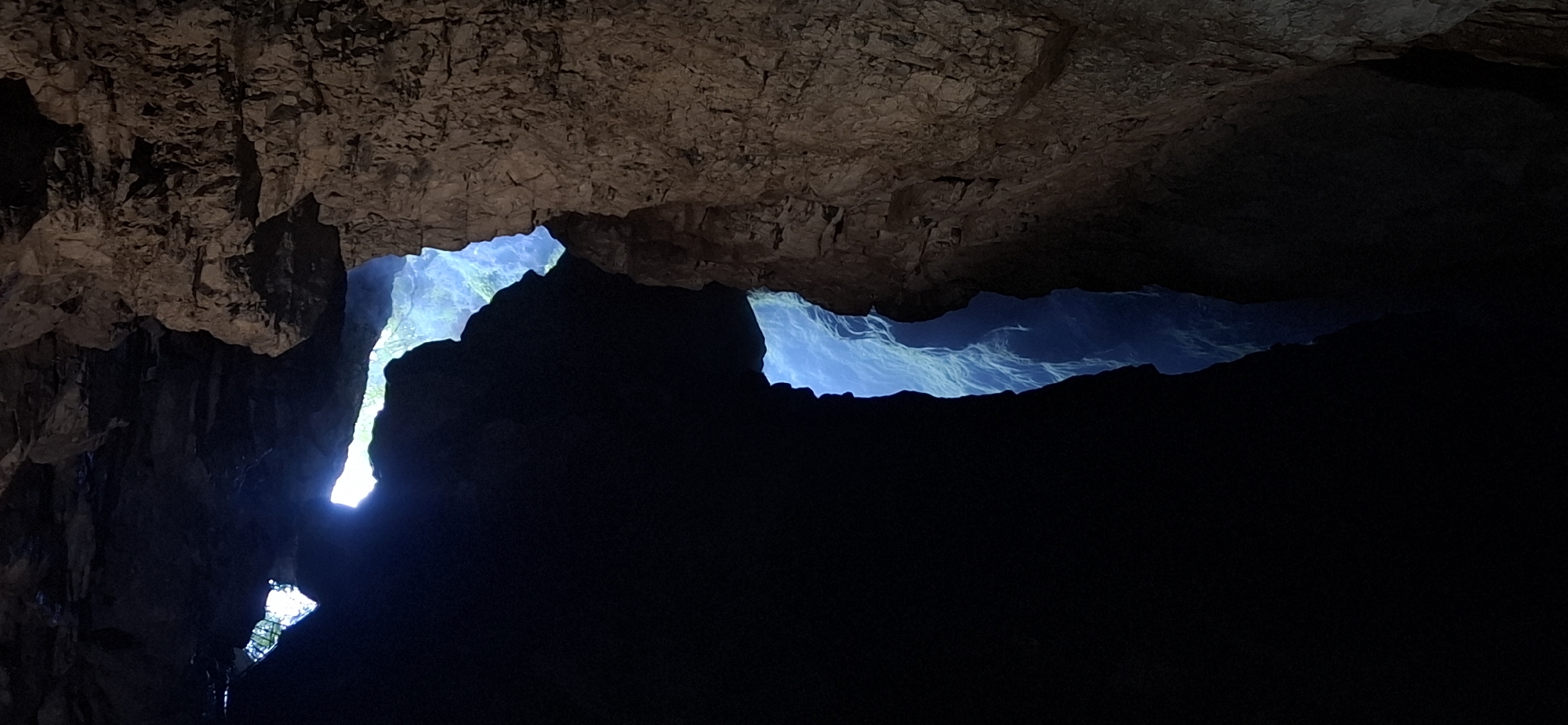 The image depicts a view from within a cave in the Trigrad Gorge, located in the Rhodope Mountains near the village of Trigrad, Bulgaria. Two openings in the cave ceiling reveal a brightly lit exterior, likely during the daytime, casting a vivid blue light into the dark interior. The foreground is dominated by the rough, textured rock formations of the cave walls and ceiling, rendered in deep shadows and earthy tones. One opening is a jagged, irregular shape, while another, further to the left, is a more horizontal fissure. Through these openings, the viewer can discern a gorge or valley below, characterized by steep, blue-tinted slopes, suggesting a significant depth and possibly a water feature or mist. The contrast between the deep darkness of the cave and the bright, ethereal light from above creates a dramatic and awe-inspiring scene.