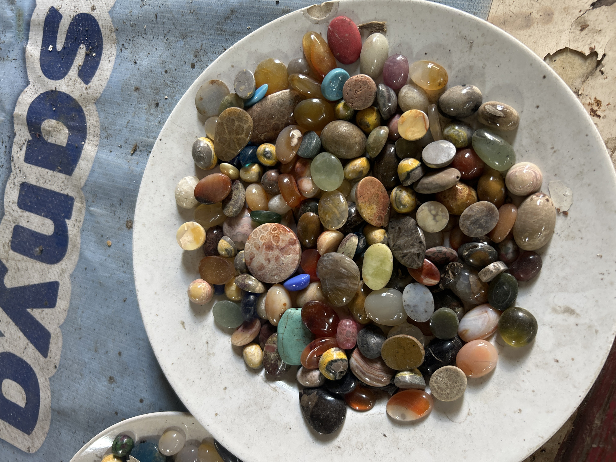 The image shows a close-up overhead view of a white plate filled with a variety of colorful, polished stones. The stones are smooth, rounded, and vary in size, shape, and color, including shades of orange, yellow, green, blue, gray, brown, pink, and white. Some stones have distinct patterns or inclusions. The plate is resting on a light blue fabric with faded blue lettering that appears to spell "BUDS". The background is a rough, textured surface with areas of peeling paint or material. The scene appears to be indoors, possibly in a workshop or a collector's display area. There are no people or actions depicted in the image. There is no information available to determine the location in Denpasar, Indonesia.