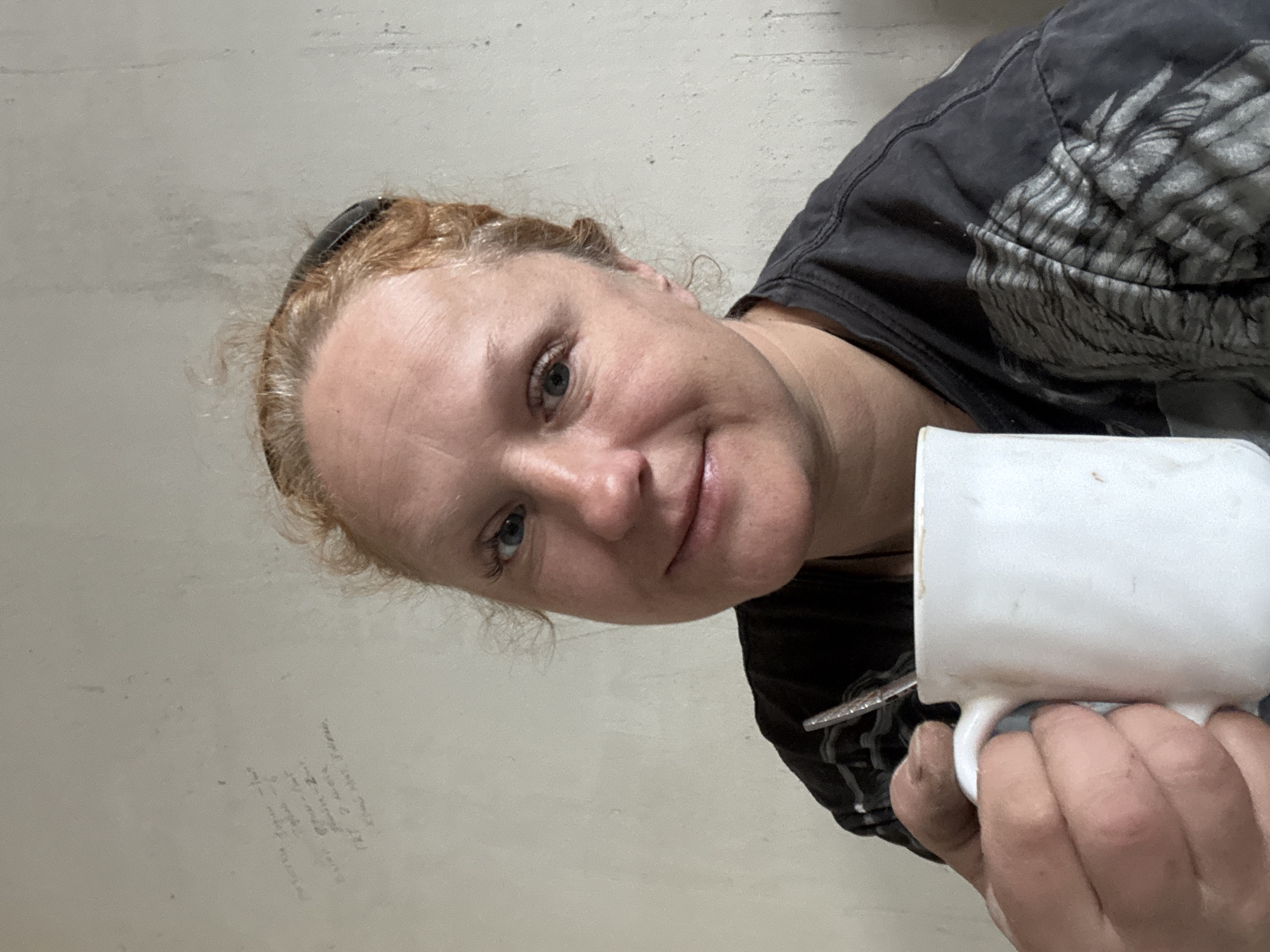 A Caucasian woman with fair skin, light brown hair, and blue eyes is holding a white ceramic mug. She is wearing a dark t-shirt. The background is a plain, light-colored wall with some faded writing. There is no identifiable information within the image to confirm the location as Chaikovsky, Russia.