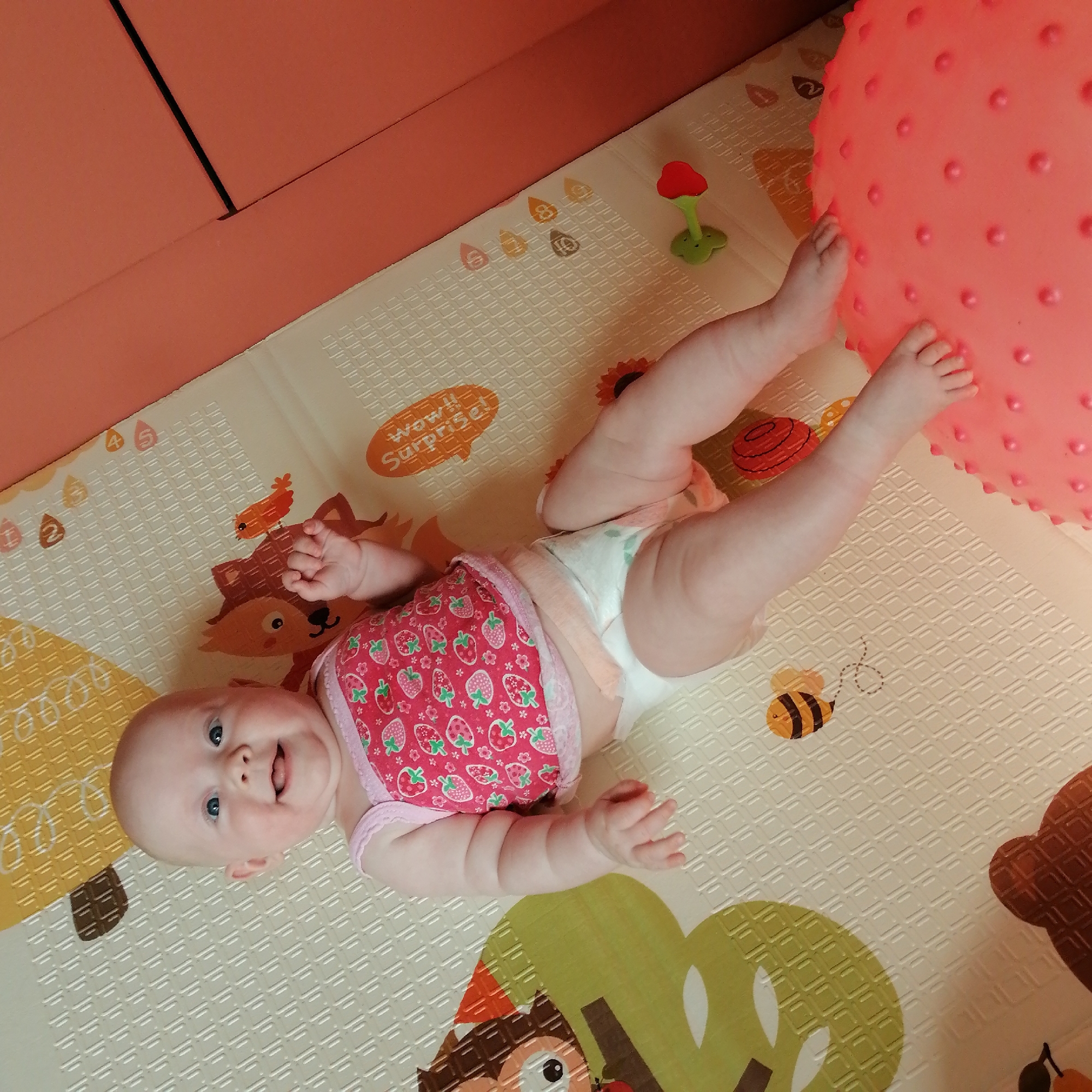 A baby girl lies on her back on a playmat. She is wearing a pink top with a strawberry pattern and a diaper. Her legs are bent and her feet are touching a large pink ball. The playmat has various colorful illustrations, including animals and a bee. The baby is looking up at the camera with a happy expression, her mouth open in a smile. The scene appears to be indoors, possibly in a home, given the playmat and the furniture visible in the background. The lighting is soft and diffused, suggesting it could be daytime. The overall mood of the image is cheerful and playful. There is no visible text in the image. The location context is Trubacheyevka, Russia.