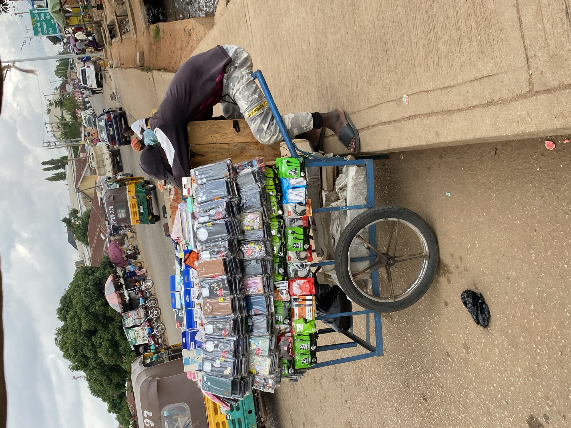 The image depicts a street scene in Bukuru, Nigeria, with a focus on a street vendor and his wares. A man, wearing a black hoodie and camouflage pants, is leaning on a wooden cart overflowing with mobile phone accessories, primarily phone cases and chargers. The cart has a blue metal frame and a large wheel. The vendor is seated on a blue metal bar attached to the cart, facing away from the camera. The cart is positioned on the side of a paved road, adjacent to a concrete sidewalk. In the background, there is traffic, including a keke (a three-wheeled motorized rickshaw), cars, and motorcycles. Several market stalls with umbrellas are visible, along with buildings and lush green trees. A signpost with place names and distances is also visible in the distance, indicating this is a thoroughfare within the city. The ground is a mix of asphalt and concrete, with some debris scattered around, including a black plastic bag. The sky is overcast.