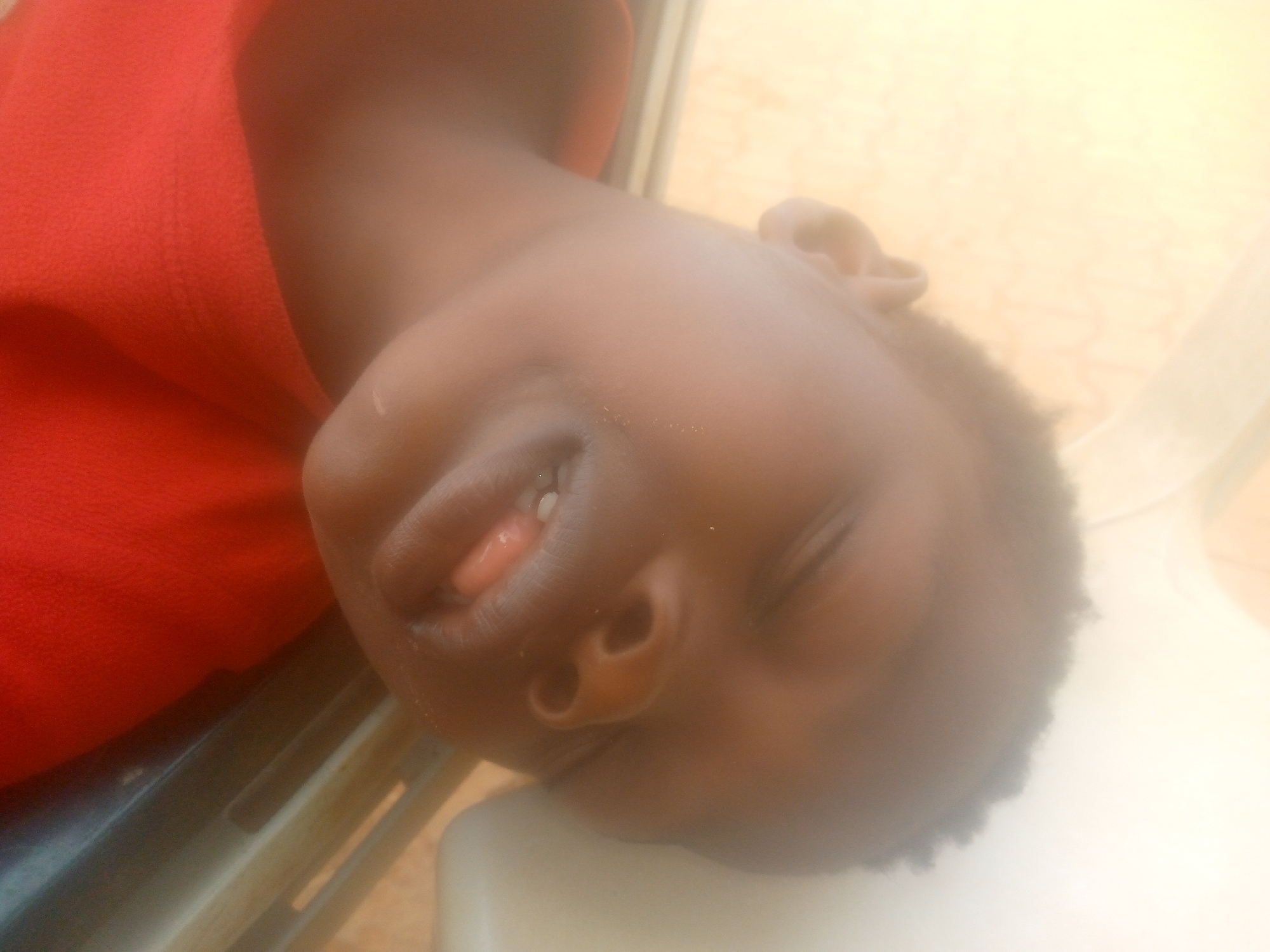 The image shows a close-up of a child, likely a young boy, with dark skin. The child is wearing a red shirt and appears to be sleeping or unconscious, with their mouth slightly open and a glimpse of their tongue visible. The child's head rests on a white surface, possibly a chair or bed. The background is blurred and indistinct, with some hints of wood grain and what appears to be a metal frame. The location is not discernible.