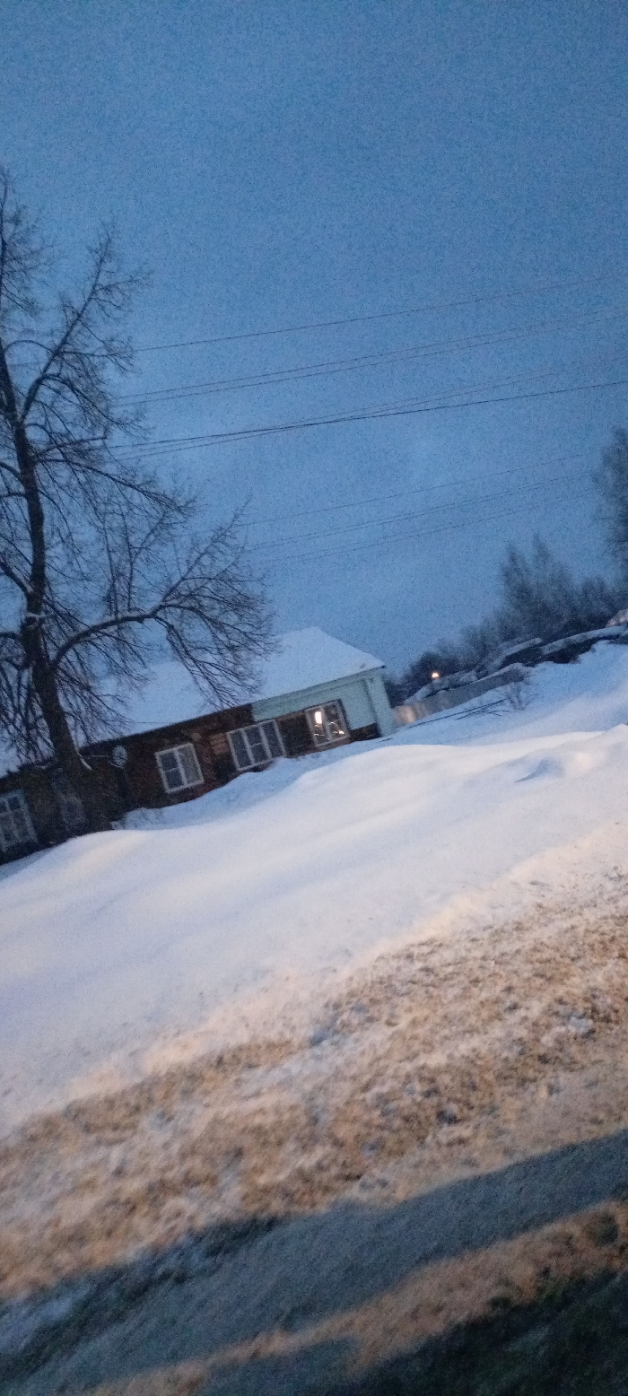 This forensic image depicts a snow-covered winter scene in Красный Маяк, Russia, under dim, blue-hued ambient light, suggestive of twilight or early dawn. The foreground and mid-ground consist of a significant snowdrift covering an uneven terrain, with patches of dark ground visible through the snow. On the left, a large, leafless deciduous tree with dark branches rises, partially obscuring the view of a building behind it. The building, located in the mid-ground, features a snow-covered pitched roof and multiple rectangular windows. Interior light emanates from at least one window on the lighter-colored section of the structure, while another section appears to be constructed of a darker material, possibly wood. Three parallel power lines traverse the upper portion of the frame against the blue sky. In the distant background, additional snow-laden trees and a faint, distant light source are visible. No individuals or discernible actions are present in the scene.
