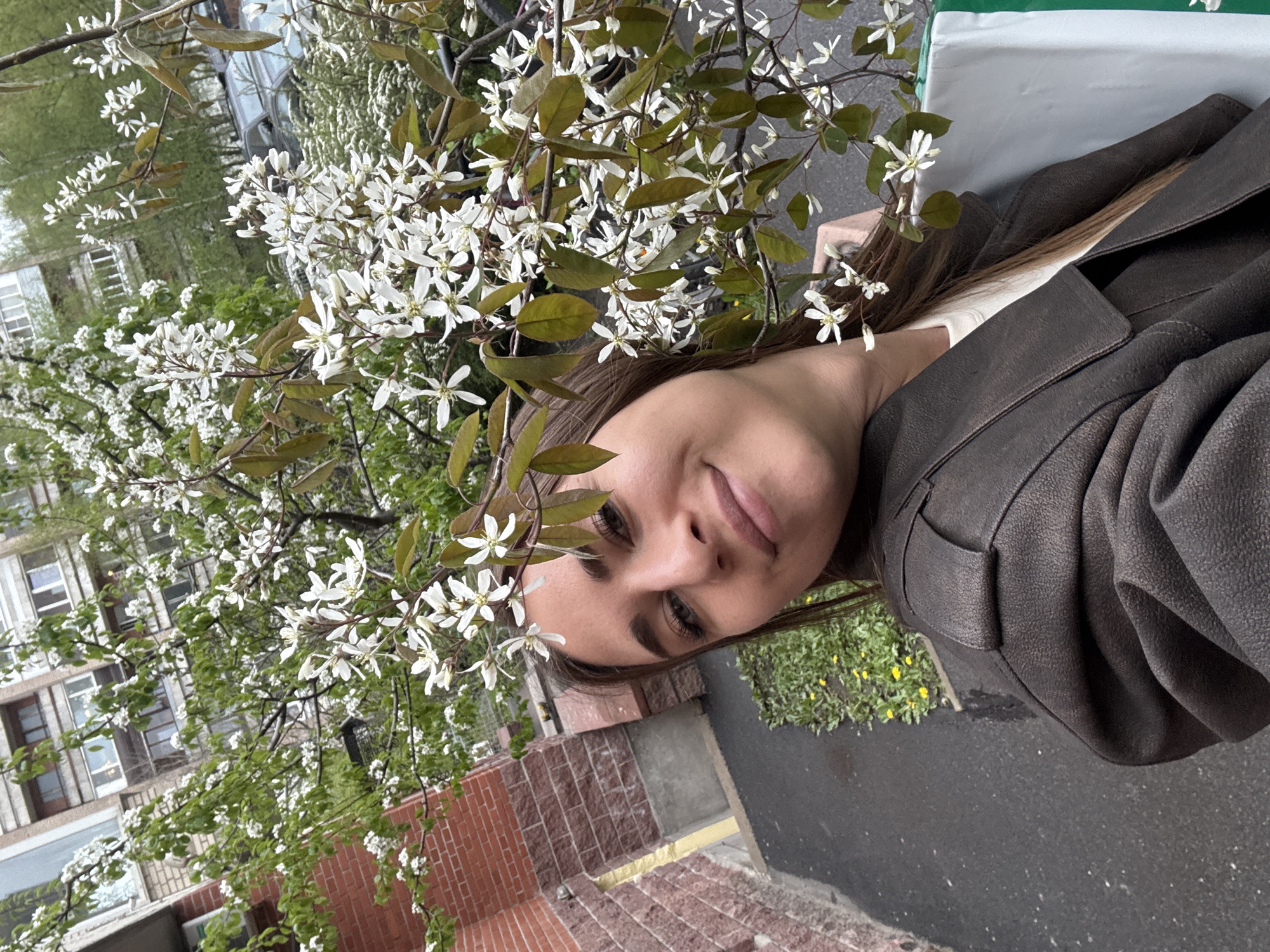 A woman with light brown hair and fair skin is outdoors next to a flowering tree with white blossoms and dark green leaves. She is wearing a brown leather jacket over a white top. The background includes a brick wall, a building with windows, greenery, and a paved surface. Based on the architectural style of the buildings and the foliage, this appears to be in Saint Petersburg, Russia.