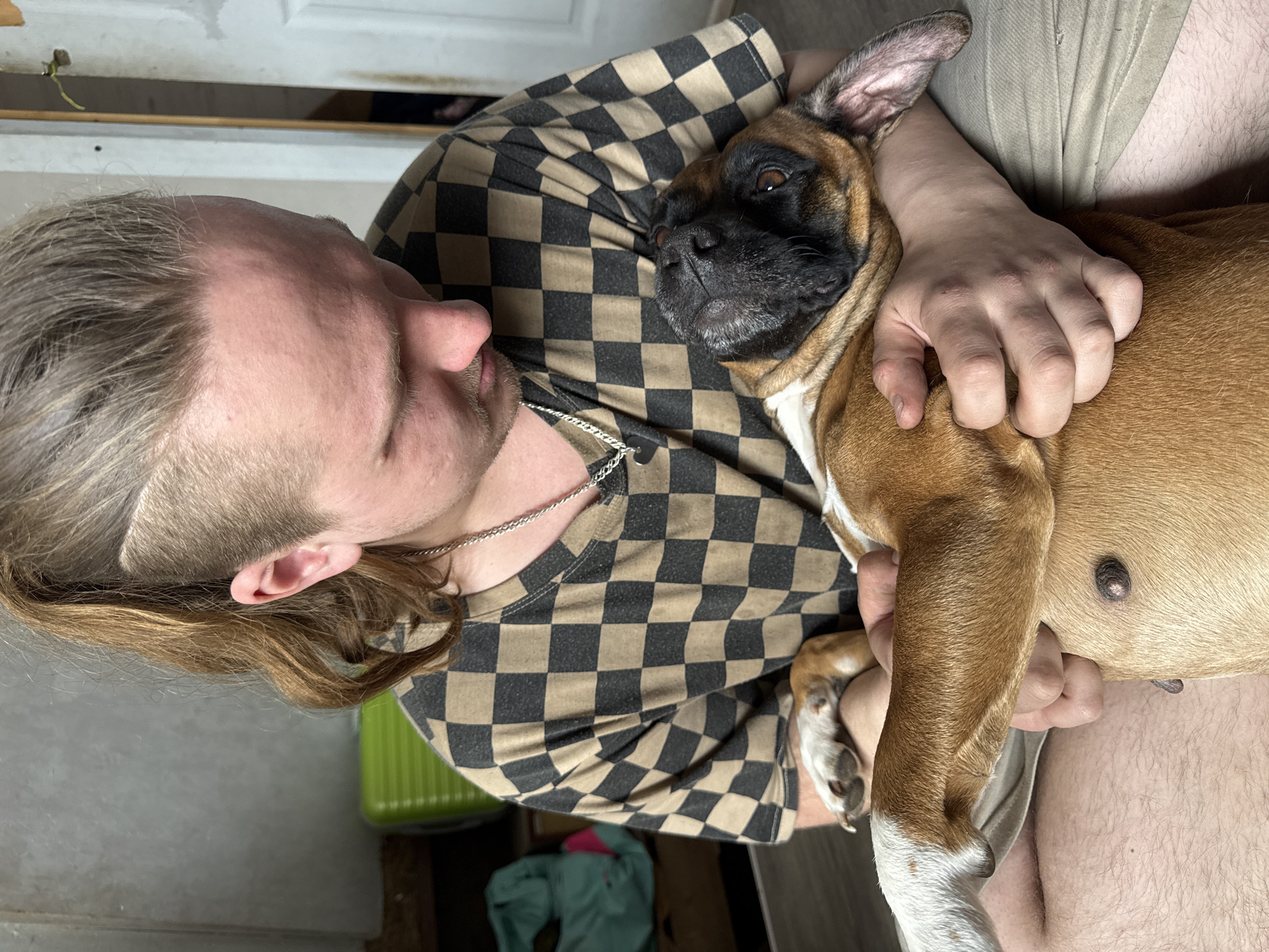 A man with long brown hair, wearing a checkered shirt, is holding a brown and black boxer dog. The man is looking at the dog, who is resting its head on his chest. The dog's front paws are resting on the man's arm. The background is blurry but appears to be indoors. The specific location in the city of Chaikovsky, Russia, cannot be determined from the image.