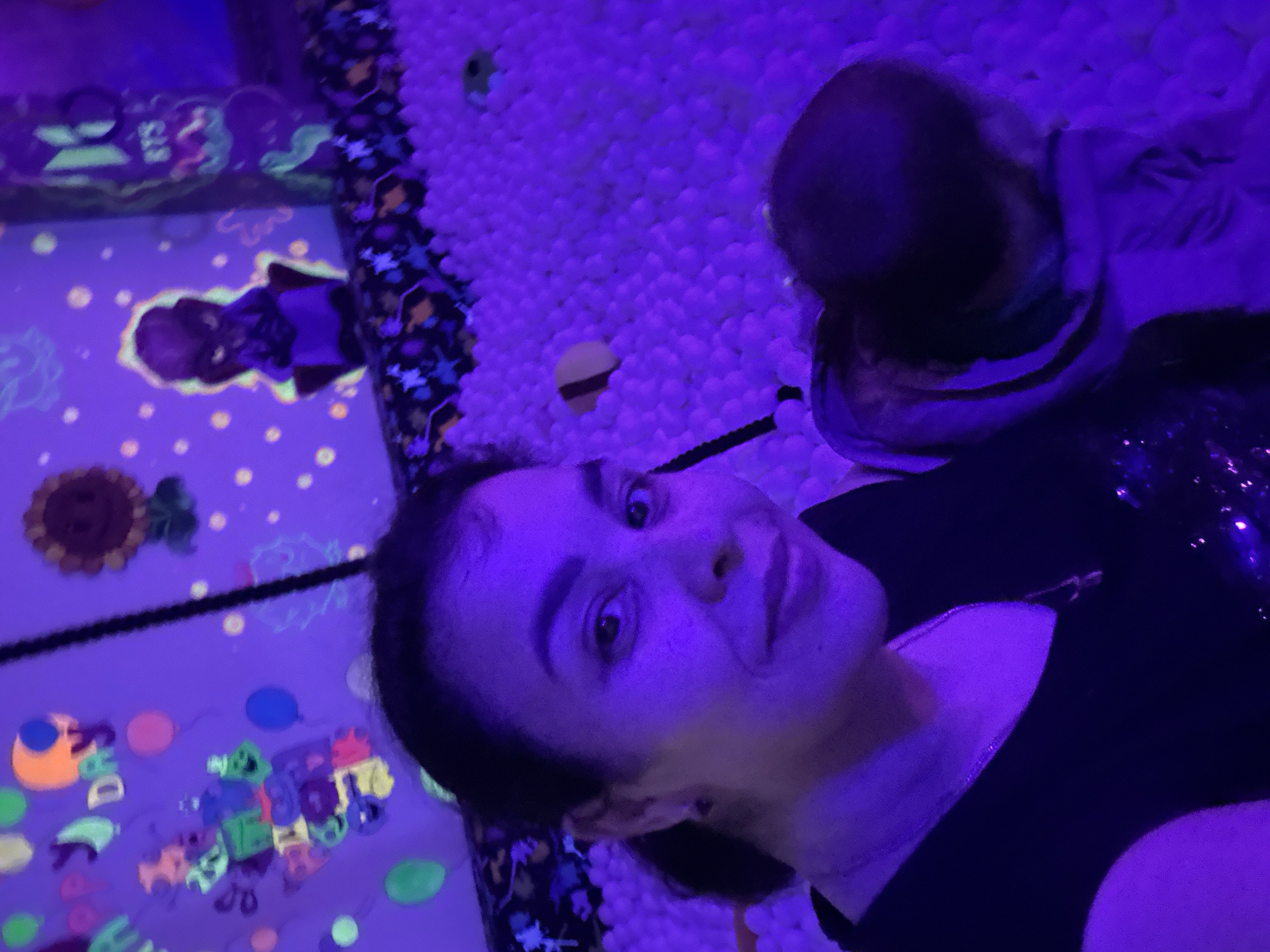 The image depicts an indoor play area, likely located in the city of Unknown, Unknown, illuminated predominantly by ultraviolet (UV) or blacklight, casting a strong purple hue over the scene. The foreground features an adult female, positioned in a large ball pit filled with countless white balls that appear luminous purple and white under the UV light. She has dark hair pulled back, visible dark eyebrows, and is looking directly at the camera with a slight smile. Her skin is tinged purple by the lighting. She wears a dark, possibly black, v-neck top with a visible metallic necklace and some sparkling embellishments on the right shoulder/chest area. A small, dark earring is visible in her right ear.
Behind the woman, the edge of the ball pit is visible, lined with a dark fabric featuring glow-in-the-dark patterns, including stars and abstract shapes in orange, yellow, and green. Further into the ball pit, to the right of the woman, a child's upper back and head are visible, facing away from the camera and slightly downwards, seemingly engaged with the balls. The child wears a light-colored hoodie or jacket. Scattered within the ball pit are a few larger, non-white objects, including a yellow, curved item and a green, rounded object.
The background consists of walls adorned with glow-in-the-dark decorations. On the left, green and yellow letters spell out parts of "HAPPY DAY" amidst various glowing balloons (blue, pink, green, orange) and other abstract or cartoon-like shapes. Toward the center-right of the background, a glowing sunflower-like character with a face and a seated, ominous-looking figure with a green outline are visible. Further to the right, a vertical panel or structure displays "BTS" in glowing green letters, surrounded by other intricate green glowing patterns. Small, glowing dots are scattered across the background walls. A dark, vertical structural element, possibly a pole or rope, extends from the top of the frame towards the woman's right shoulder.