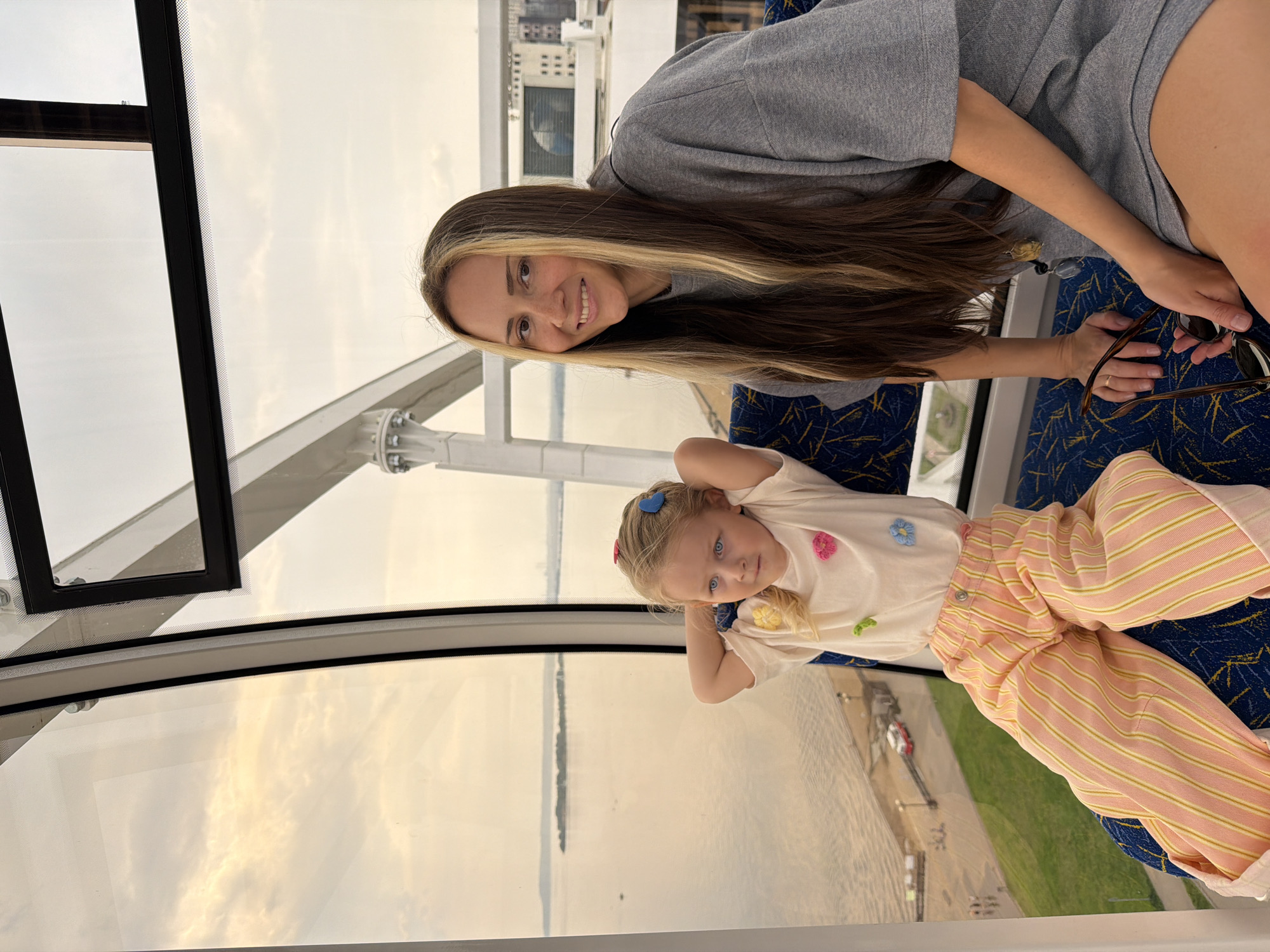 The image shows a woman and a young child inside a cabin, seemingly on a Ferris wheel. The woman has long, brown hair with blonde highlights and is wearing a grey top. She is smiling at the camera and holding a pair of sunglasses. The child has blonde hair with blue hair clips and is wearing a white shirt with decorative flower accents and striped pink and yellow pants. She is looking at the camera with her hands behind her head. The cabin has blue patterned seats. Through the window, a view of Saratov, Russia's river, land, and cityscape can be seen. The day appears to be overcast.