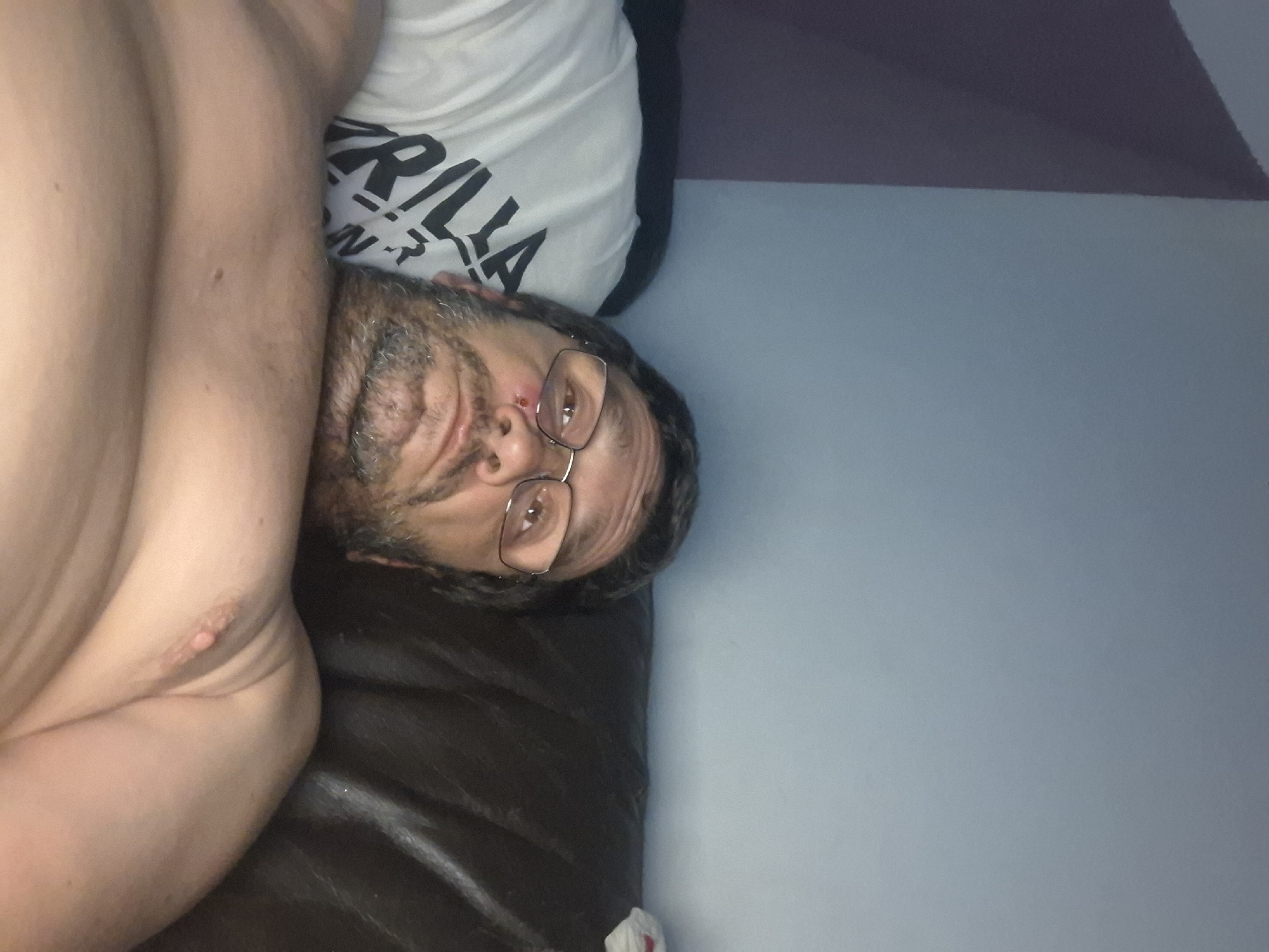An undated, low-angle, close-up, indoor selfie shows a man lying on a dark brown leather couch. The man is facing upwards towards the camera and is wearing glasses. He has short, dark hair, a beard, and a scrape or bruise on his left cheekbone. He is shirtless, and a white t-shirt with the word "GORILLA" printed in black lettering is visible under his left arm. The background consists of a light blue wall with a darker colored wall to the left. There are no visible signs of the city of Yerocham, Israel, in this image.