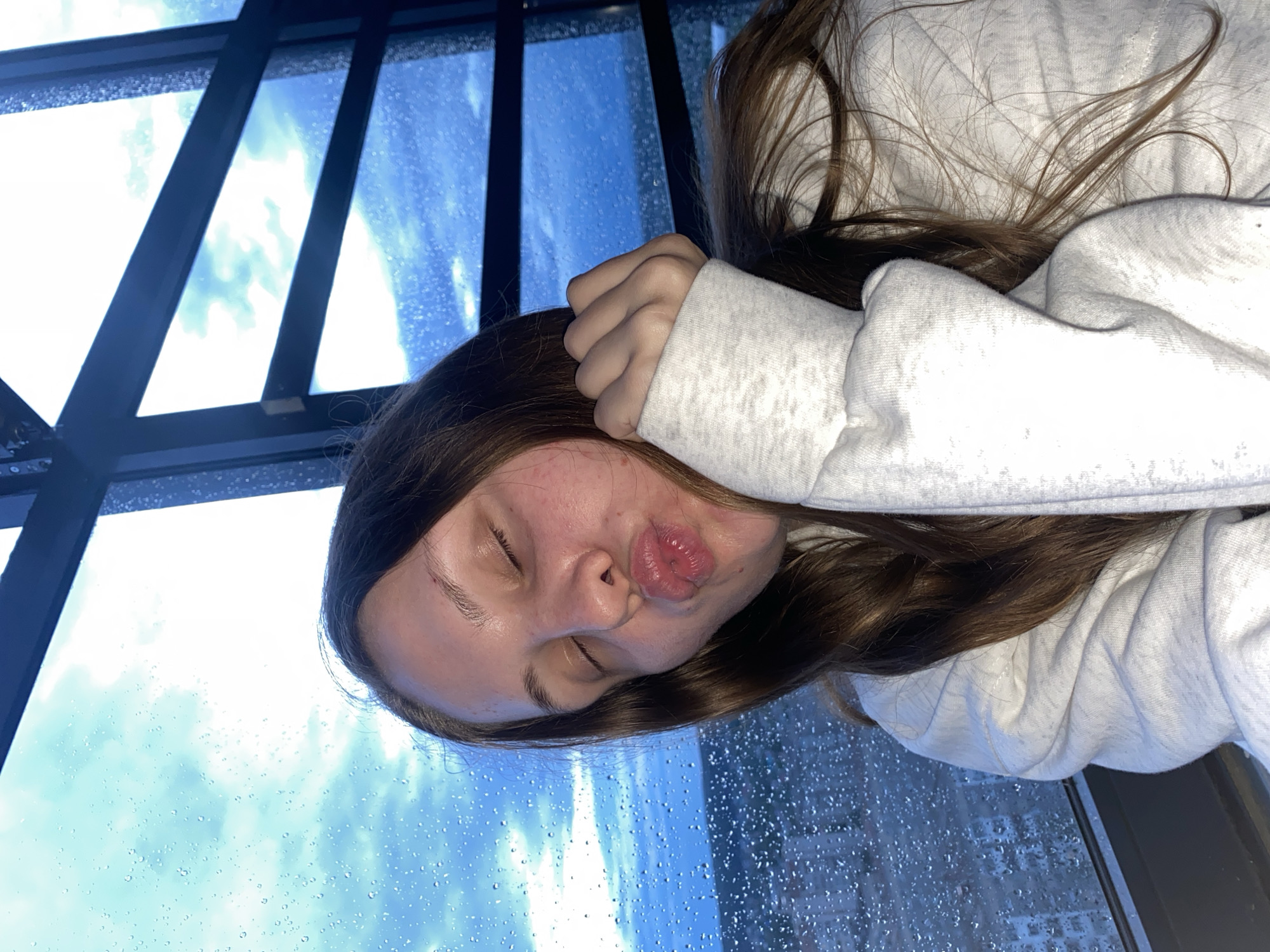 The image shows a young woman with her eyes closed and her lips puckered, as if blowing a kiss. She is wearing a white hoodie and her hand is resting on her cheek. She is sitting in front of a large window, through which a city skyline can be seen. The window is covered in raindrops, suggesting that it is raining. The sky is cloudy and the light is dim, indicating that it is either early morning or late evening. The overall mood of the image is one of quiet intimacy and reflection.