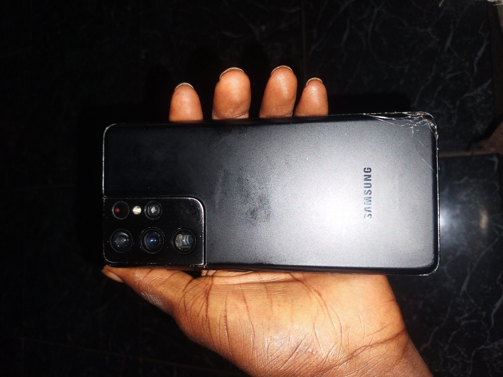 A black Samsung smartphone with a cracked upper right corner is held in a dark-skinned hand against a dark background. The word "SAMSUNG" is printed vertically on the back of the phone. The phone has multiple camera lenses on the left side. The location is Jos, Nigeria.