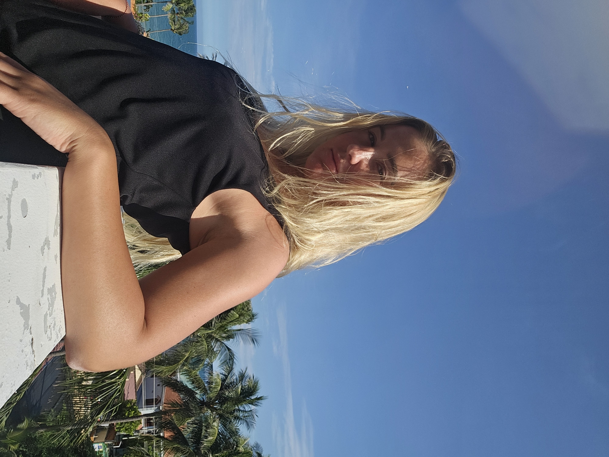 A young woman with blonde hair is leaning on a railing with a tropical landscape in the background. The sky is clear and blue. She is wearing a black tank top and looking at the camera with a neutral expression. The ocean and palm trees are visible in the distance, suggesting a coastal location, possibly in Phu Thuy Ward, Vietnam.