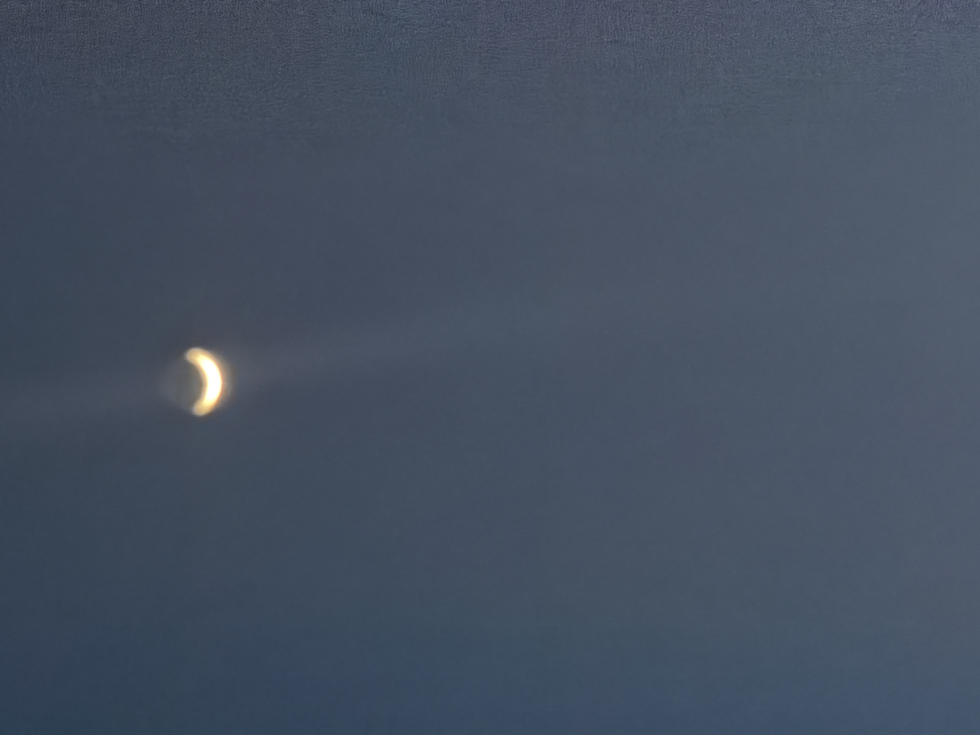 The image shows a hazy crescent shape, likely the sun during a solar eclipse, against a dark blue sky. The crescent is bright yellow and out of focus, with a faint halo of light surrounding it. The sky appears textured, possibly due to atmospheric conditions or the camera's sensor. There are no people, objects, or environmental features visible besides the sky and the celestial phenomenon. The location in Bukuru, Nigeria, is an assumption based on provided context and is not visually confirmed by the image itself.