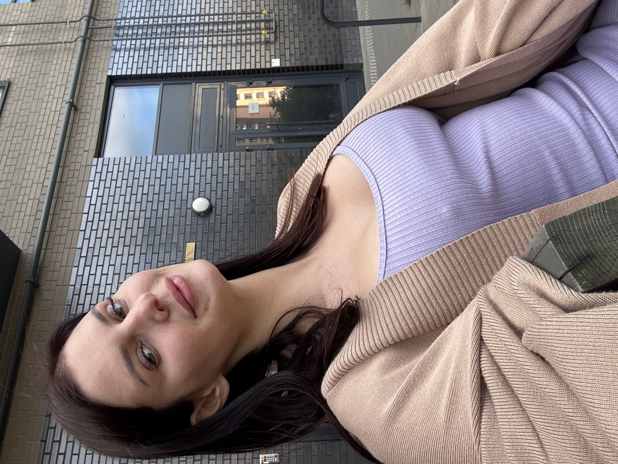 A medium-shot, vertical, outdoor photo captures a young woman with long, dark hair and a fair complexion. She is wearing a lilac-colored ribbed tank top and a light beige cardigan. Her head is tilted, and she is looking directly at the camera with a soft expression, her lips slightly parted and her eyes appearing to be a shade of green. Her makeup includes blush on her cheeks and eyeliner, and her eyebrows are well-defined.
The background consists of a modern building with a facade of grey and beige bricks. A dark-colored window is visible, reflecting the sky and a distant building with multiple floors and windows. Pipes run vertically along the building. The overall lighting suggests it might be daytime, possibly overcast due to the soft lighting and lack of harsh shadows. The woman appears to be seated, as indicated by the portion of a wooden bench or railing visible in the lower right of the frame. There is no discernible text in the image.