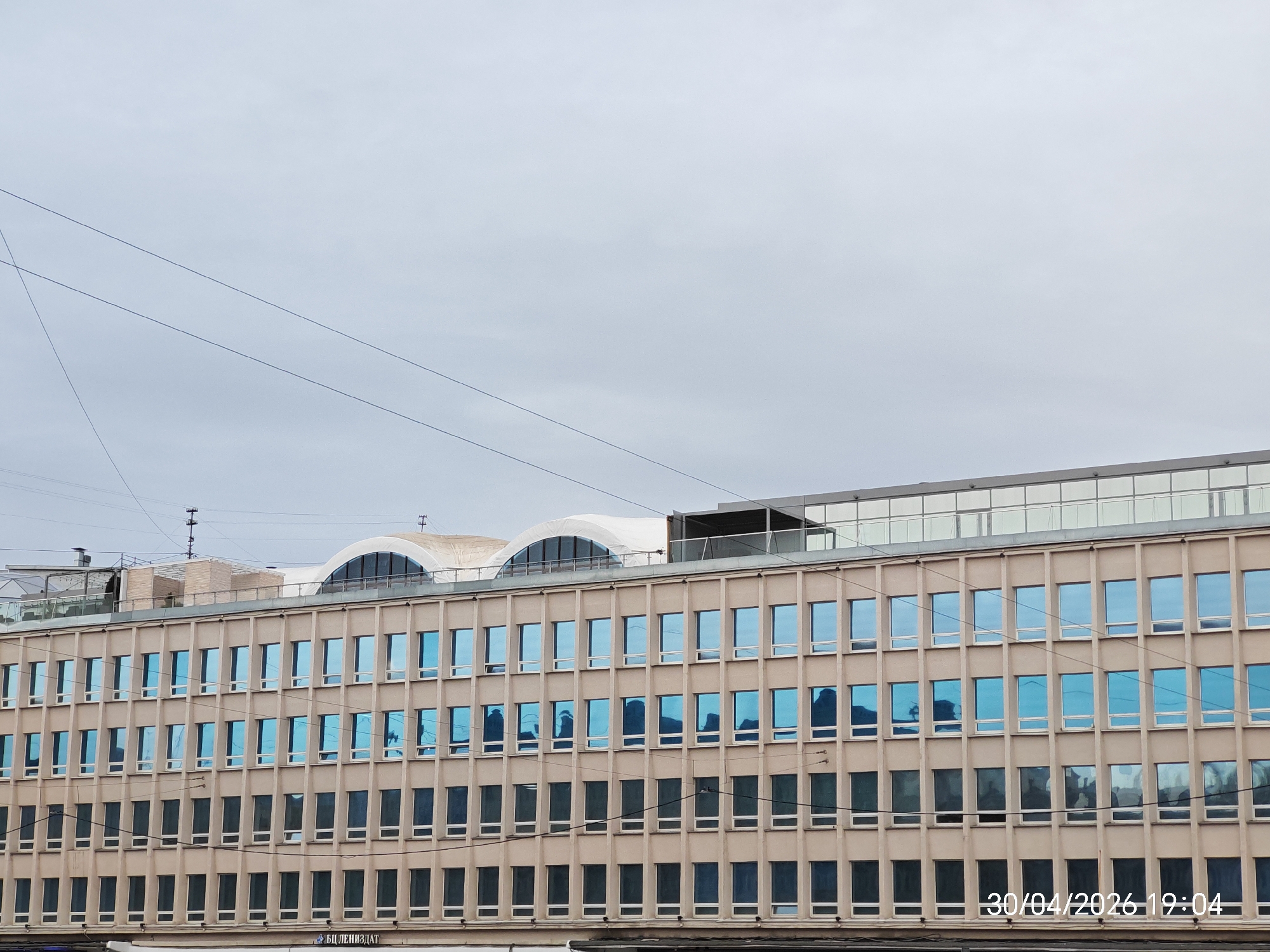 An eye-level shot captures a large, multi-story building under an overcast sky, with power lines crisscrossing the upper portion of the frame. The building features a beige, grid-like facade with numerous rectangular windows arranged in regular rows and columns. The upper three visible floors have windows that strongly reflect the blue sky, giving them a distinct blue tint and showing indistinct reflections of other structures or elements. The windows on the lower visible floors appear darker, possibly due to tinting or interior reflections.
Above the main facade, the building's roofline reveals modern architectural elements, including two large, arched, white structures that could be skylights or decorative roof features, and a flat section with what appears to be glass railings or walls. Various antennae or ventilation systems are visible on the rooftop. Dark power lines run horizontally across the building's facade in the middle section.
The sky is a uniform light grey, indicating an overcast day. Several thin, dark power lines stretch diagonally across the upper left and middle sections of the sky. In the bottom right corner of the image, a timestamp reads "30/04/2026 19:04". Faint Cyrillic text, "БЦ ЛЕНИЗДАТ", is visible at the bottom of the building's facade, suggesting a business or building name. The overall scene depicts a functional, likely commercial or office building in an urban environment during what appears to be early evening on a cloudy day.