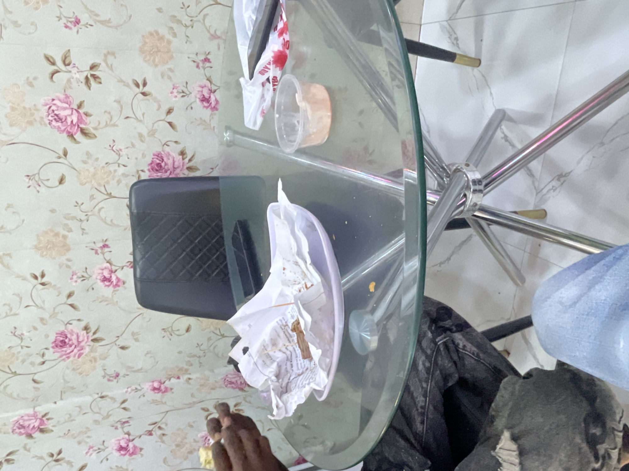 In a room in Jalingo, Nigeria, a circular glass table with a chrome base sits on a tiled floor. The table has a floral wallpaper backdrop. On the table, there is a white plate with crumpled white paper that appears to have remnants of food, suggesting a meal has recently been eaten. To the right, a small clear plastic container holds a light orange substance, possibly a sauce or dip. A black plastic bag with red lettering is also visible on the table. A black chair with a quilted pattern is positioned behind the table. In the foreground, a person's hands, holding a piece of food, are visible on the left side of the frame, and their legs, clad in denim, are in the lower part of the image. The overall scene depicts a casual dining setting, likely after a meal.