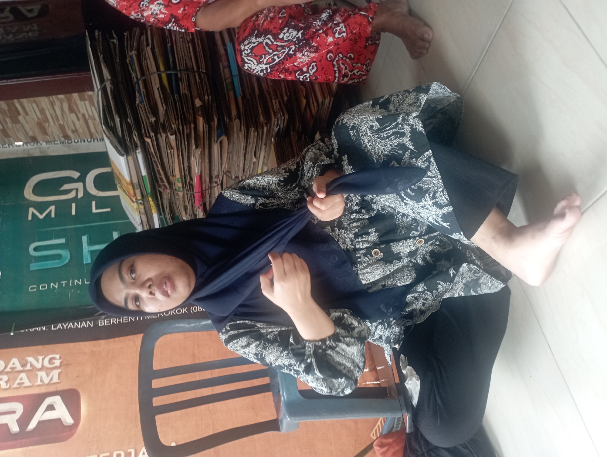 In a dimly lit indoor setting, likely a room in Maos Kidul, Indonesia, a woman wearing a dark blue hijab and a black and white patterned dress is seated on the floor. Her left hand is seen holding a piece of the dark blue fabric. She is looking towards the camera with a neutral expression. To her right, another person, partially visible, is wearing a red and white patterned skirt and also sitting on the floor. The background features a stack of cardboard boxes and a dark plastic chair. The floor appears to be tiled.