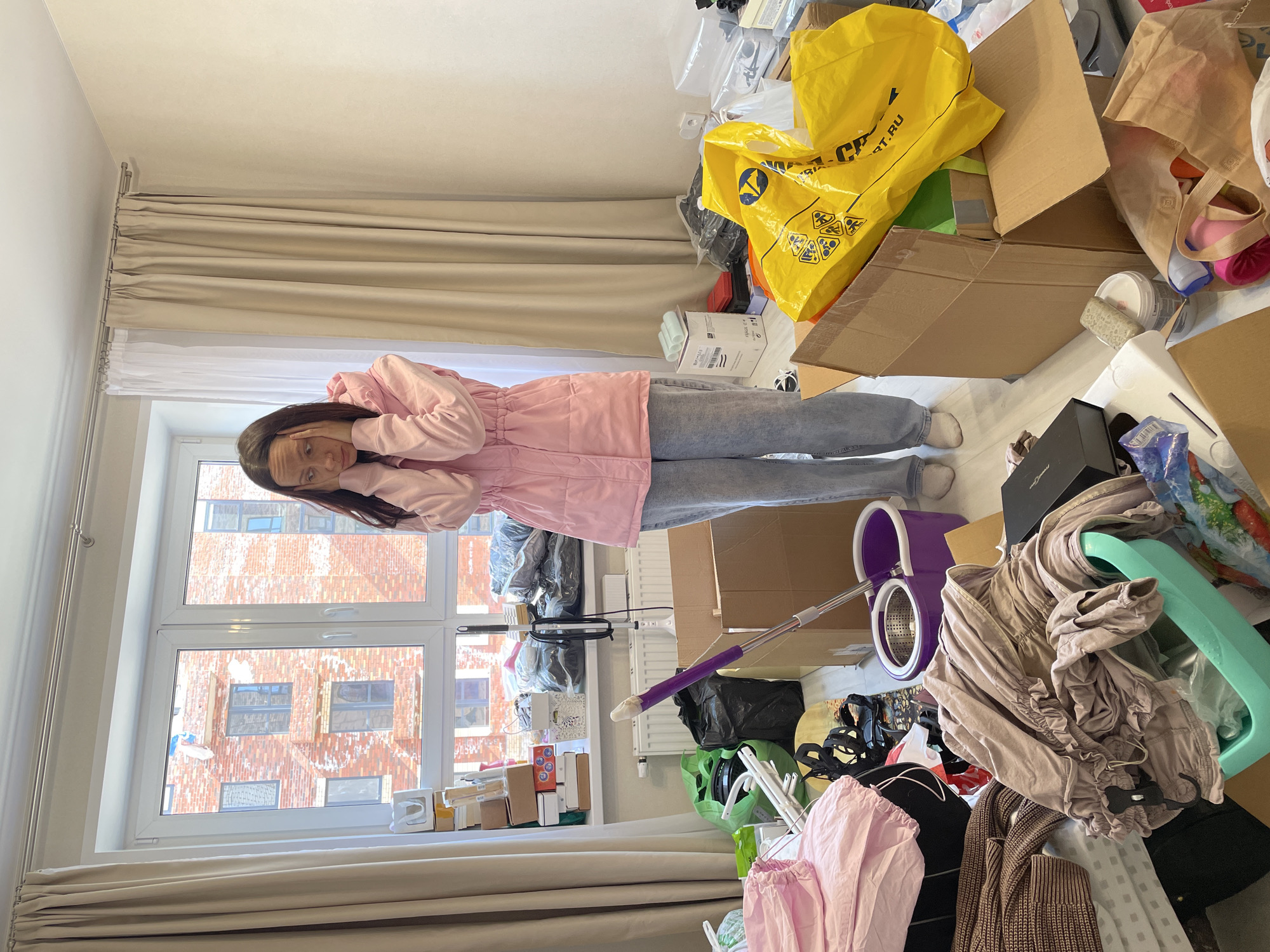 An indoor scene depicts a cluttered room, possibly in Izhevsk, Russia, with a young woman standing near a large window, appearing overwhelmed. She has long dark hair and is wearing a light pink hooded jacket or windbreaker, light blue jeans, and light-colored socks or slippers. Her hands are up to her face, partially covering it, and her expression suggests dismay, stress, or exhaustion.
The room is filled with numerous items, creating a sense of disarray. To the right of the woman and extending throughout the room are stacks of cardboard boxes, many open, and various plastic bags, including large bright yellow ones. One yellow bag clearly displays the text "ART.RU" along with several triangular warning symbols. Piles of clothes, some neatly folded but mostly scattered, are visible, alongside household items like a purple spin mop bucket and a green plastic basin. Black trash bags are also present. The floor appears to be light-colored wood or laminate.
A large multi-pane window dominates the background, revealing the exterior of red-brick buildings with multiple windows, characteristic of an urban environment. Bright daylight streams through the window, indicating it is daytime. Light beige pleated curtains and white sheer blinds cover the top portion of the window. The walls and ceiling are a plain, light color. The overall impression is of a room undergoing a chaotic move, unpacking, or major clear-out, with the woman feeling stressed by the situation.
