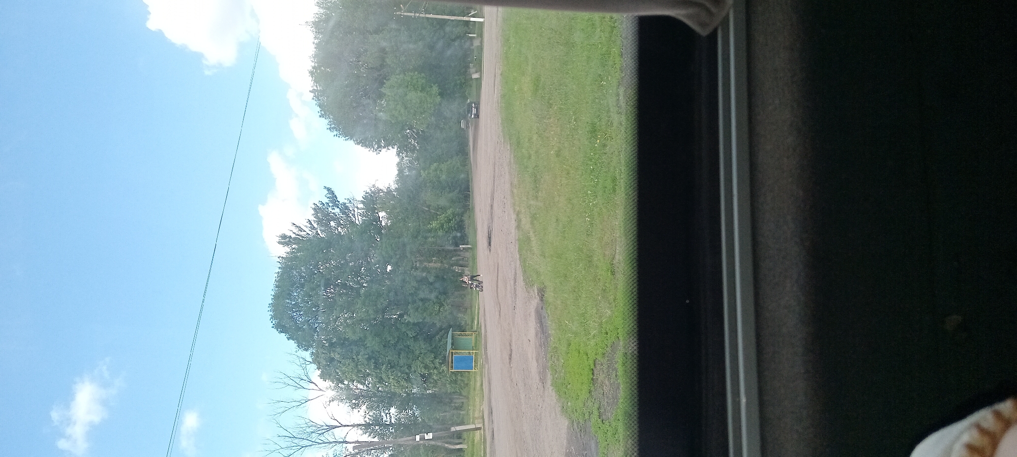 A person pushes a stroller on a dirt road in front of a small bus stop shelter with a blue and yellow roof. The shelter is located in front of a large deciduous tree. Behind the trees, a clear blue sky with scattered white clouds can be seen. There are two vehicles visible in the distance. The foreground shows a portion of a vehicle's interior, possibly a car window, with a beige curtain on the right side. The scene appears to be in a rural or suburban area.