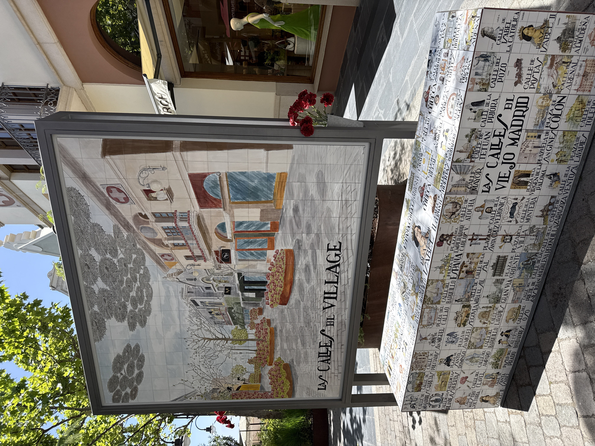 The image shows a large tiled mural depicting a street scene with buildings, trees, and flowers. Below the mural is a tiled bench that also features various images and text. The mural and bench are set outdoors on a paved area, with trees and buildings visible in the background. The overall impression is of a public space decorated with artistic tile work.
The mural is rectangular and mounted on a metal frame. The scene depicted is a European-style street with multi-story buildings, balconies with flowers, and cobblestone pavement. The sky above the buildings is light blue with stylized grey clouds. The bench in front of the mural is also rectangular and covered in tiles. These tiles display a collage of images, portraits, and text, many of which appear to be street names and historical figures or scenes related to Madrid.
The environment suggests a bright, sunny day. Sunlight casts shadows on the paved ground. In the background, a shop window displays a mannequin in a green dress.
While the mural itself seems to be a general depiction of a street, the bench specifically references "LAS CALLES DEL VIEJO MADRID," indicating a connection to the old city of Madrid. The text on the bench includes various street names from Madrid such as "PLAZA DEL SOL", "CALLE MAYOR", "GRAN VIA", and "CALLE DE ALCALA", and the style suggests it might be located in a shopping or pedestrian area. The location is likely a tourist area or a public square in a city that celebrates its history and architecture through art. The specific mention of Las Matas-pinar-monte Rozas in the prompt suggests this artwork may be located there, possibly as a representation of Madrid's streets within that locale, or as part of a larger public art initiative.
