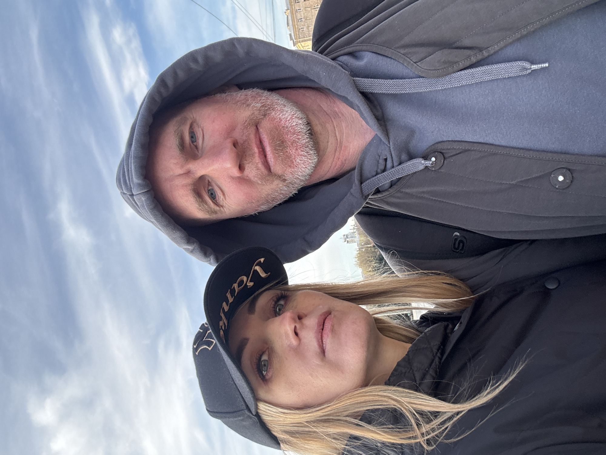 A man and a woman are in a selfie. The man, with fair skin, blue eyes, and a gray beard, wears a gray hoodie pulled over his head and a dark jacket. The woman, with blonde hair, fair skin, and green eyes, wears a black baseball cap with "Yankees" embroidered in gold lettering, along with a black jacket. The sky is cloudy with visible light. The location appears to be Saint Petersburg, Russia, judging by the city's architectural style in the background.