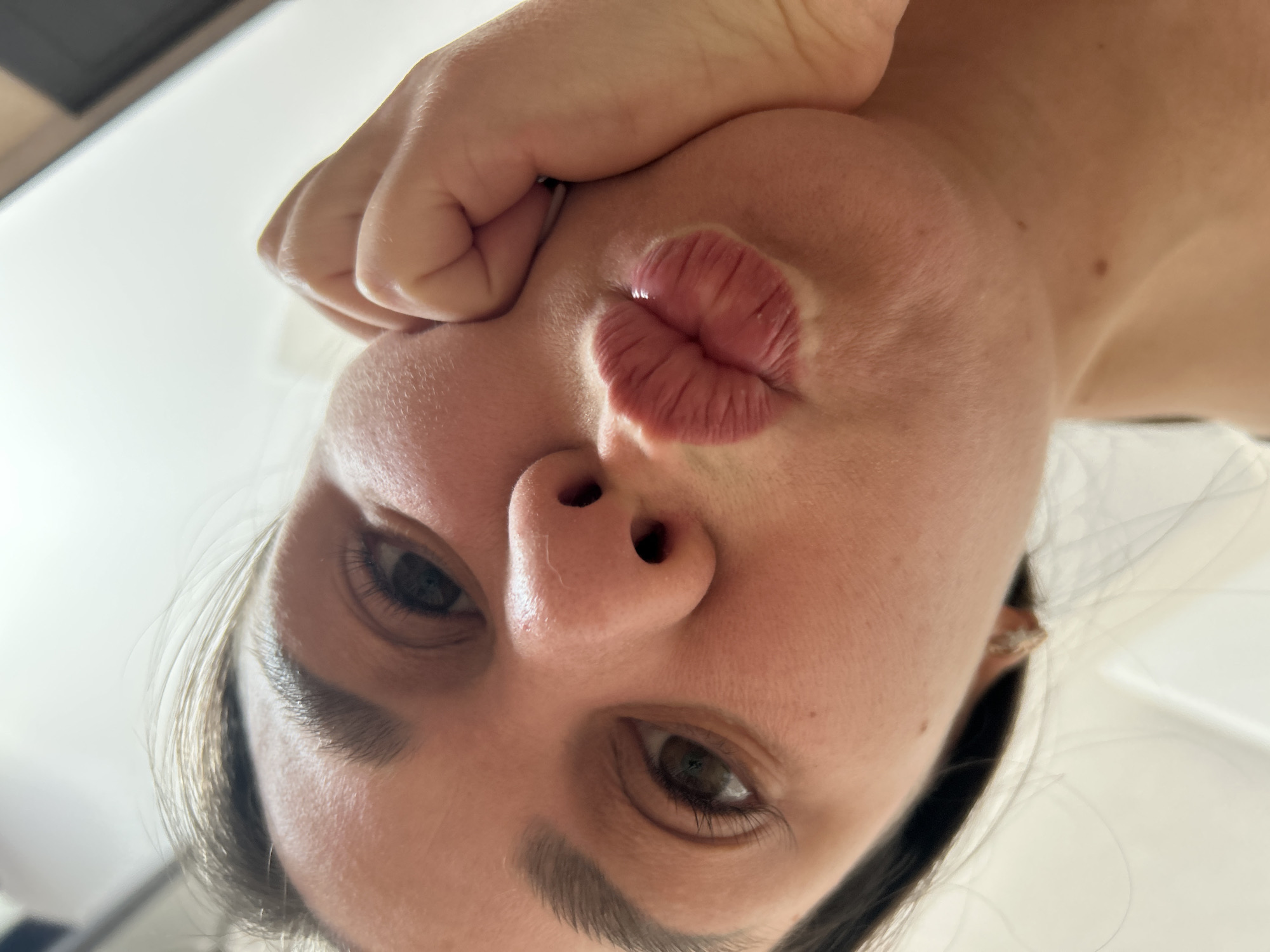 A close-up, tilted selfie shows a woman's face from below. She is making a "duck face" with her lips pursed. Her right hand is clenched into a fist and pressed against her right cheek. She has brown eyes and eyebrows. Her hair is dark and pulled back. The background is a mix of white and a dark undefined object. There is no information to support any location in the city of Chaikovsky, Russia.