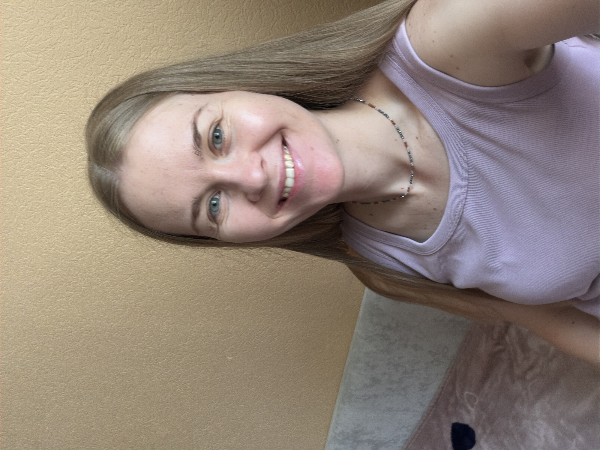 A medium shot captures a blonde woman in her late 20s or early 30s smiling at the camera. She has blue eyes and light skin, and her long, straight blonde hair is parted in the middle. She is wearing a light purple tank top and a beaded necklace. The background is a textured, light brown wall and a portion of a sofa with a pink and white patterned blanket. There are no identifiable landmarks or contextual clues that would indicate the location in Khabarovsk, Russia.