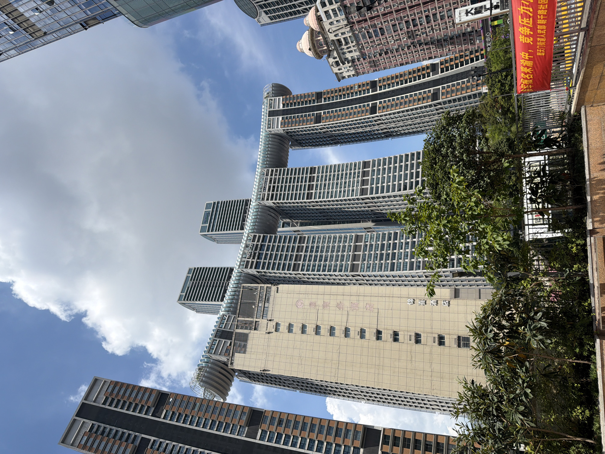 A low-angle, wide shot captures the cityscape of Yuzhong District, China, under a partly cloudy blue sky. The dominant subjects are several towering modern skyscrapers, including the distinctive "Crystal" building with its horizontal element connecting two towers. The Crystal building, in the center, features a beige facade on its main structure with a series of regularly spaced windows. Red vertical Chinese characters are visible on this beige section, identifying it as "锦江酒店" (Jinjiang Hotel). To the left of the Crystal building is another tall, dark-colored skyscraper with many windows. To the right, a colorful, ornate building with dome-like structures is visible, with a modern skyscraper to its right. In the foreground, lush green trees and bushes are planted behind a metal fence. A red banner with white Chinese text is also visible in the lower right portion of the image, near the fence. The overall scene depicts a dense urban environment with striking contemporary architecture.