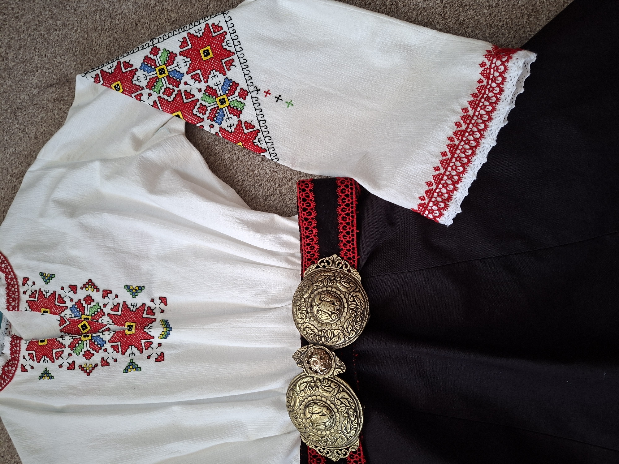 A close-up, overhead shot shows details of a white, embroidered folk costume. The costume includes a white blouse with colorful geometric embroidery around the neckline and sleeves. The sleeves are long and end with white lace trim with red embroidery. A black skirt is worn with a wide, decorative belt. The belt has two ornate, gold-colored buckles and is adorned with red lace detailing. The background is a textured brown carpet.