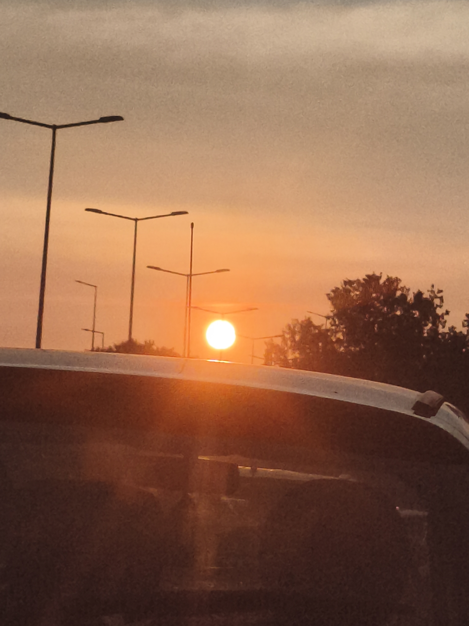 The image depicts a sunset scene viewed from inside a vehicle, likely on a road or bridge, in Nowy Dwór Mazowiecki, Poland. The sun, a bright orb of light, is positioned low in the sky, casting a warm orange glow that diffuses through the atmospheric haze. Several streetlights, silhouetted against the sky, are visible, suggesting an urban or suburban setting. The foreground is dominated by the curved surface of a car's roof or windshield, reflecting the ambient light and the surrounding environment. Trees are visible in the middle ground on the right side of the frame, partially obscuring the horizon. The sky is a gradient of soft orange and muted gray tones.